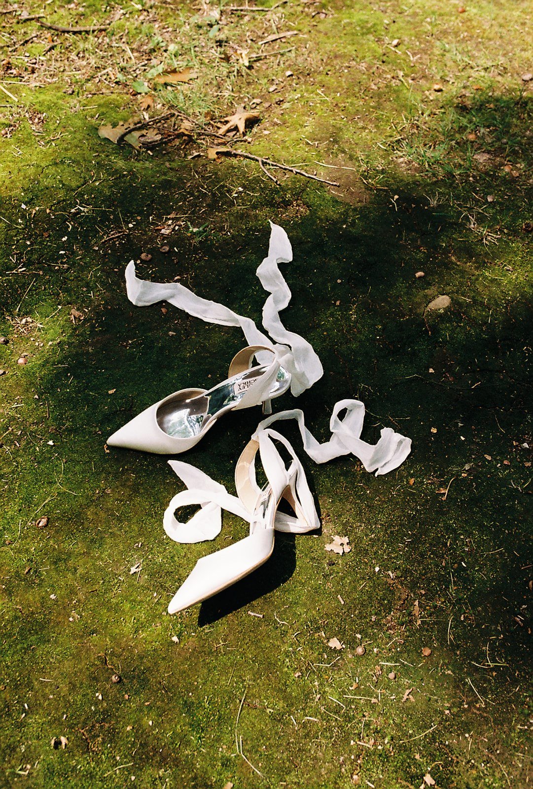 A pair of white high-heeled shoes with tissue paper inside is on a moss-covered ground outdoors, with some dried leaves and small twigs scattered around.