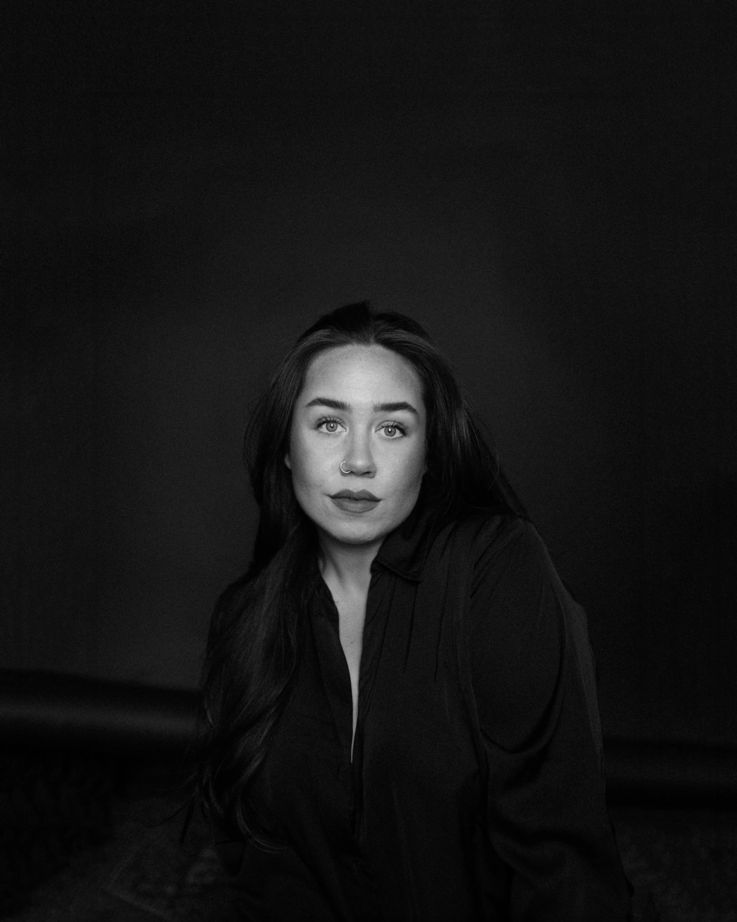 A woman with light skin, dark hair, and blue eyes looking directly at the camera, with a glittering nose ring, against a plain dark background.