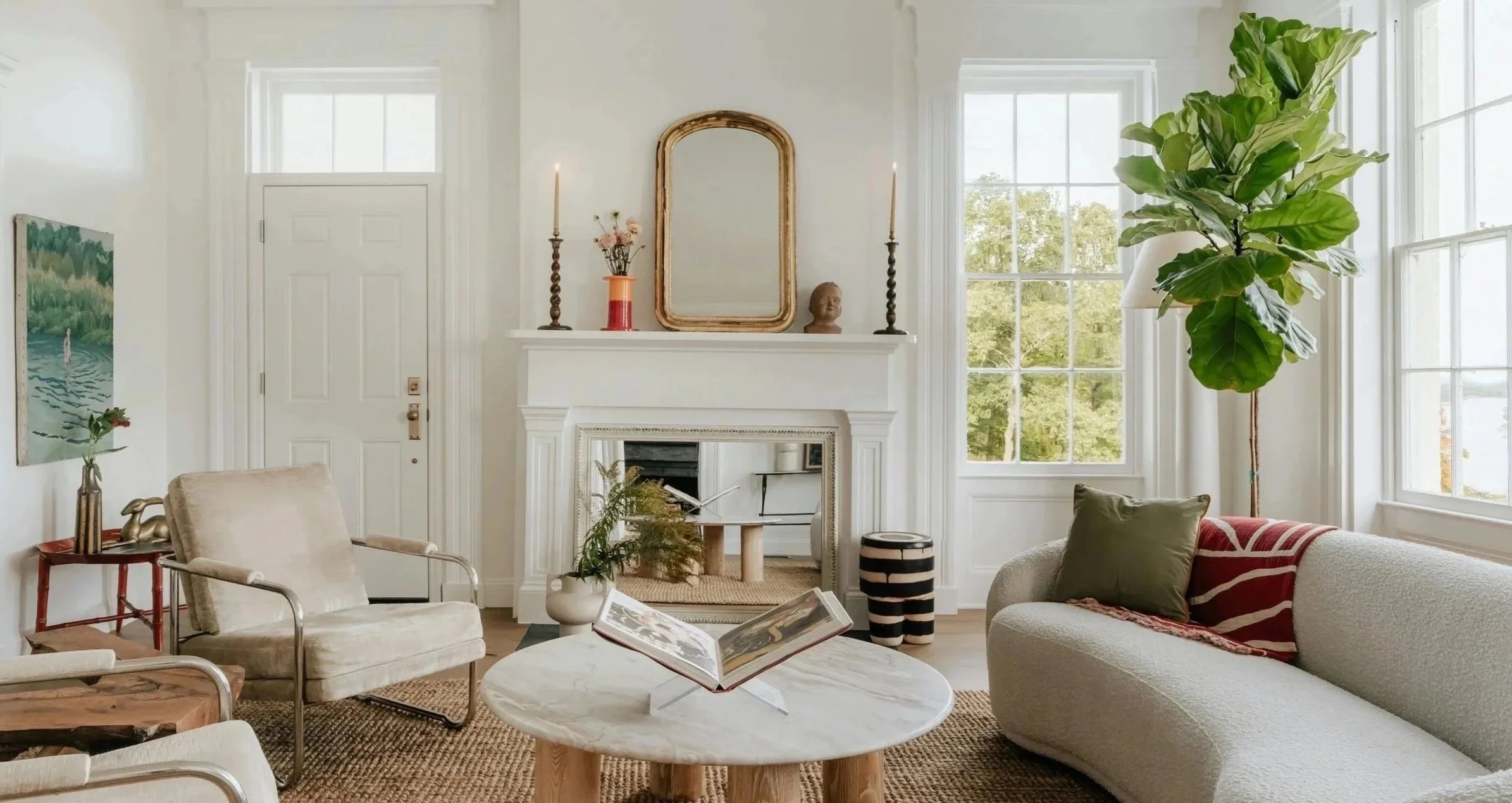 Cozy living room with white walls, large windows, a white fireplace with decor, a tall fiddle leaf fig plant, and vintage furniture including a beige armchair and a rounded cream sofa with a green pillow and a patterned throw, accented by artwork and decorative objects.