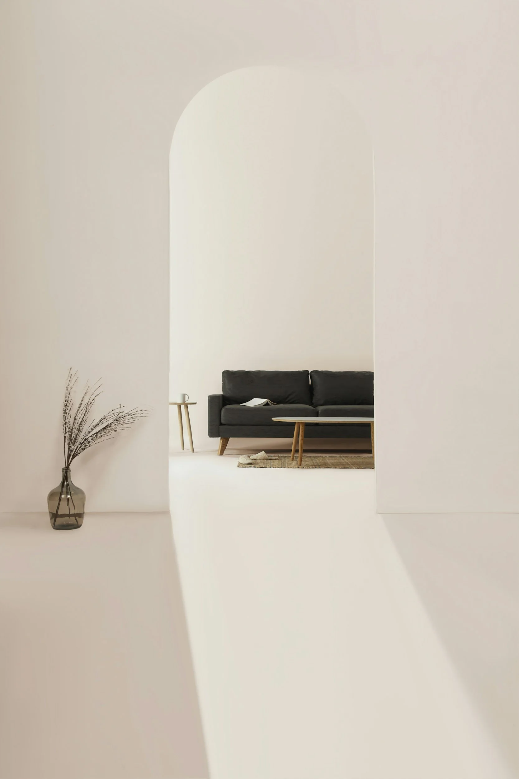 View through an arched doorway to a living room with a black sofa, a wooden coffee table, and a small side table with a book, against white walls and a light-colored floor.