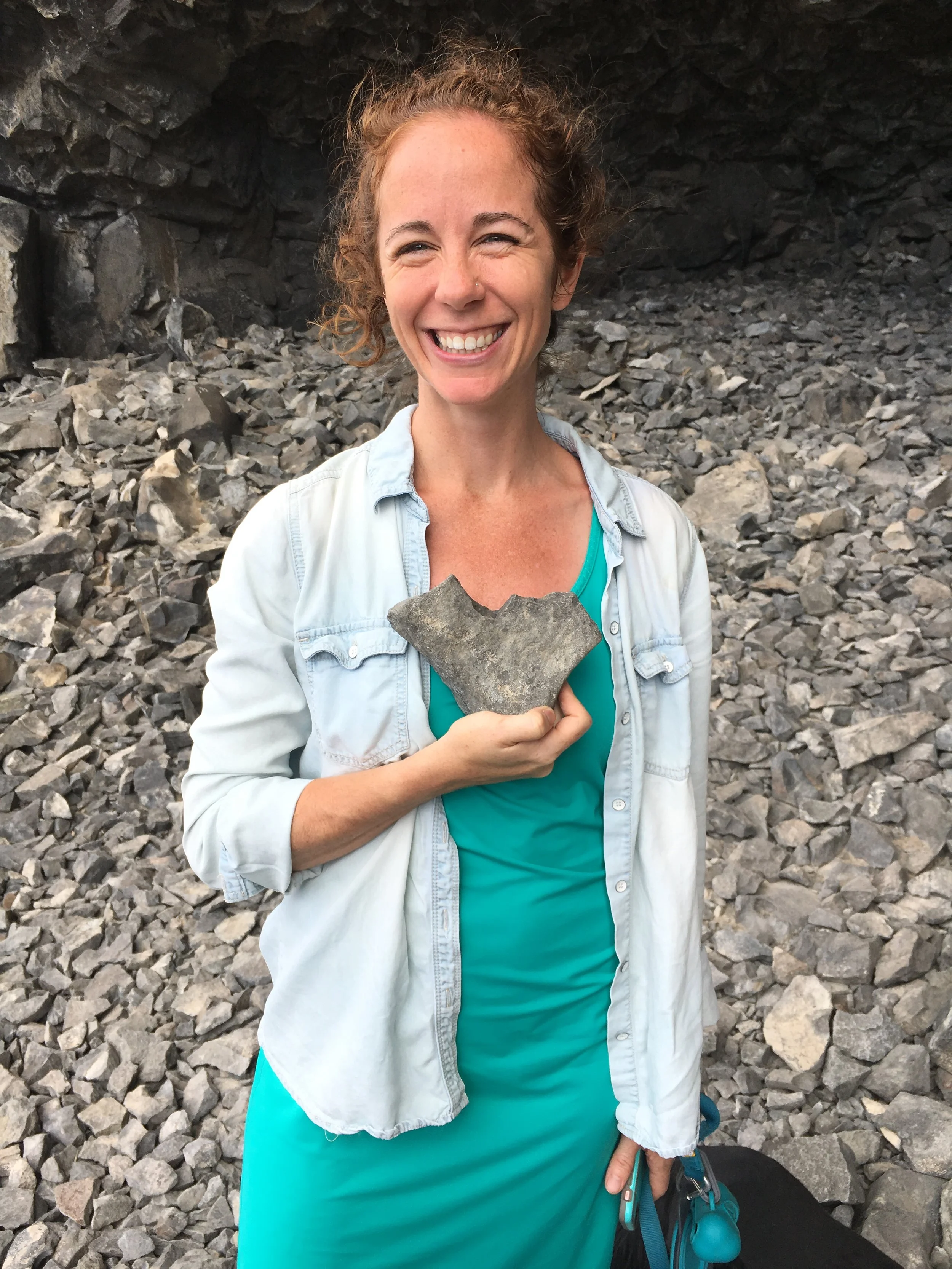 Erinn Zavaglia,  is smiling while holding a rock, standing on rocky terrain in front of a stone cave or wall.