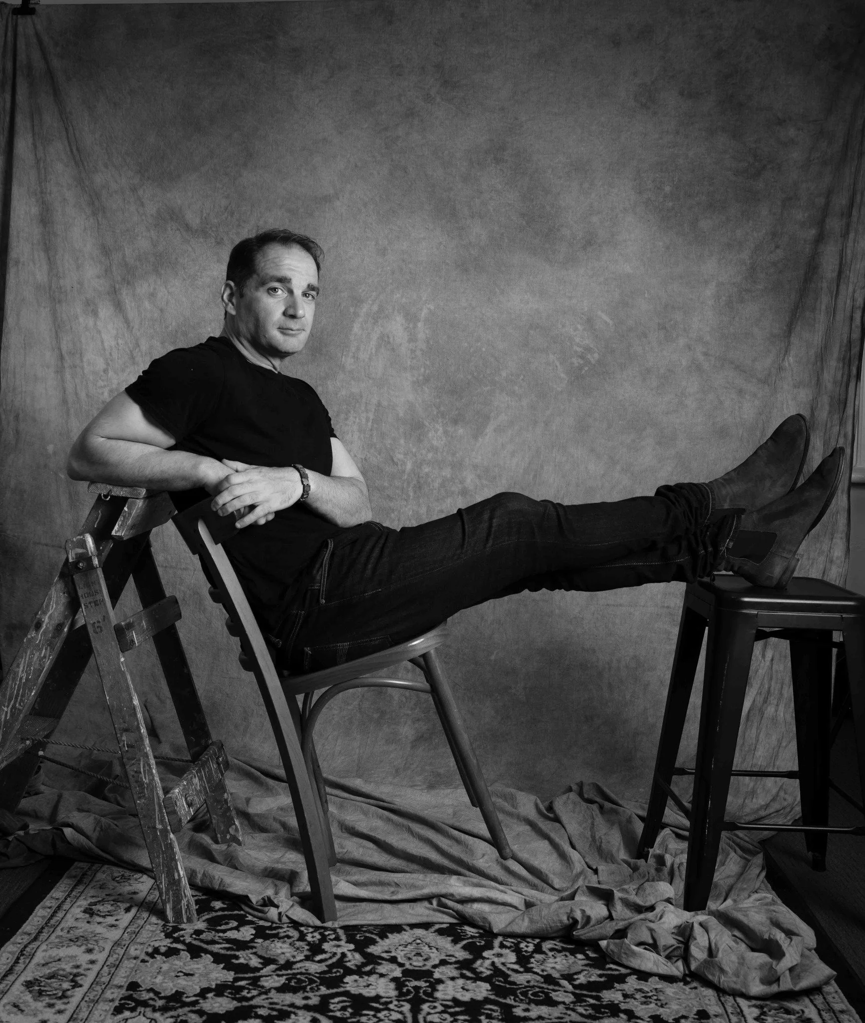A black and white photo of a man reclining on a wooden ladder with his feet elevated on a chair, against a textured backdrop with a patterned rug on the floor and crumpled paper underneath.