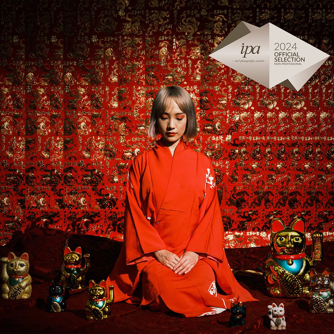 A woman dressed in a red kimono sitting on a dark red bed with various gold and colorful Maneki-neko figurines surrounding her, against a red patterned background with gold accents.