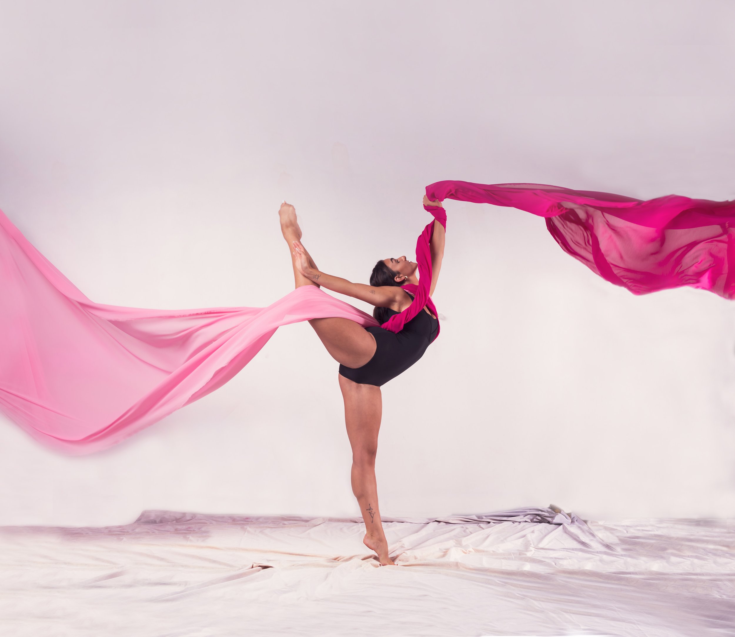 A woman performs a balancing dance pose holding pink and magenta flowing fabrics against a plain white background.