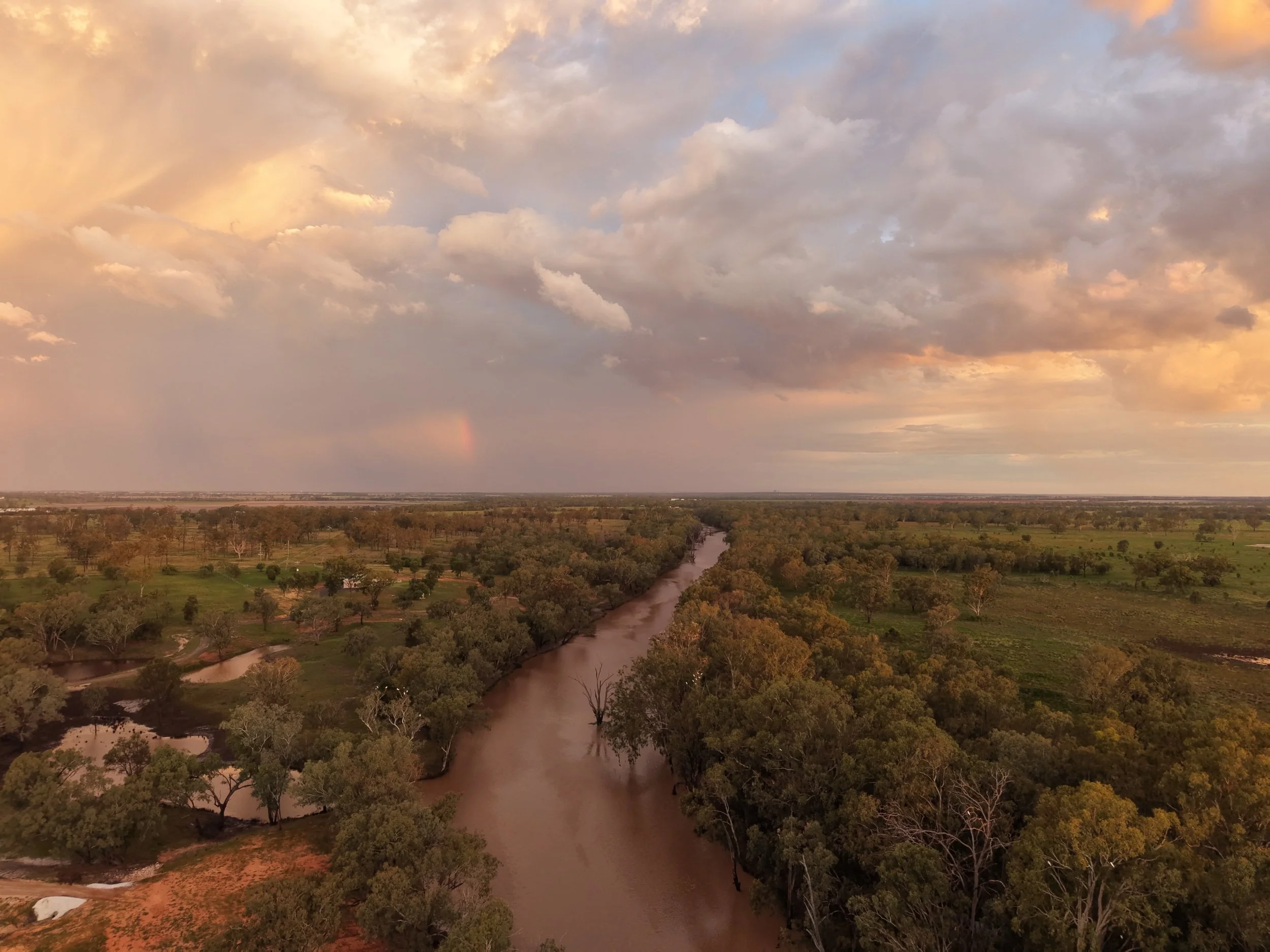 A wide view of a river flowing through a landscape with scattered trees, under a partly cloudy sky with warm, muted tones and a faint rainbow in the distance.