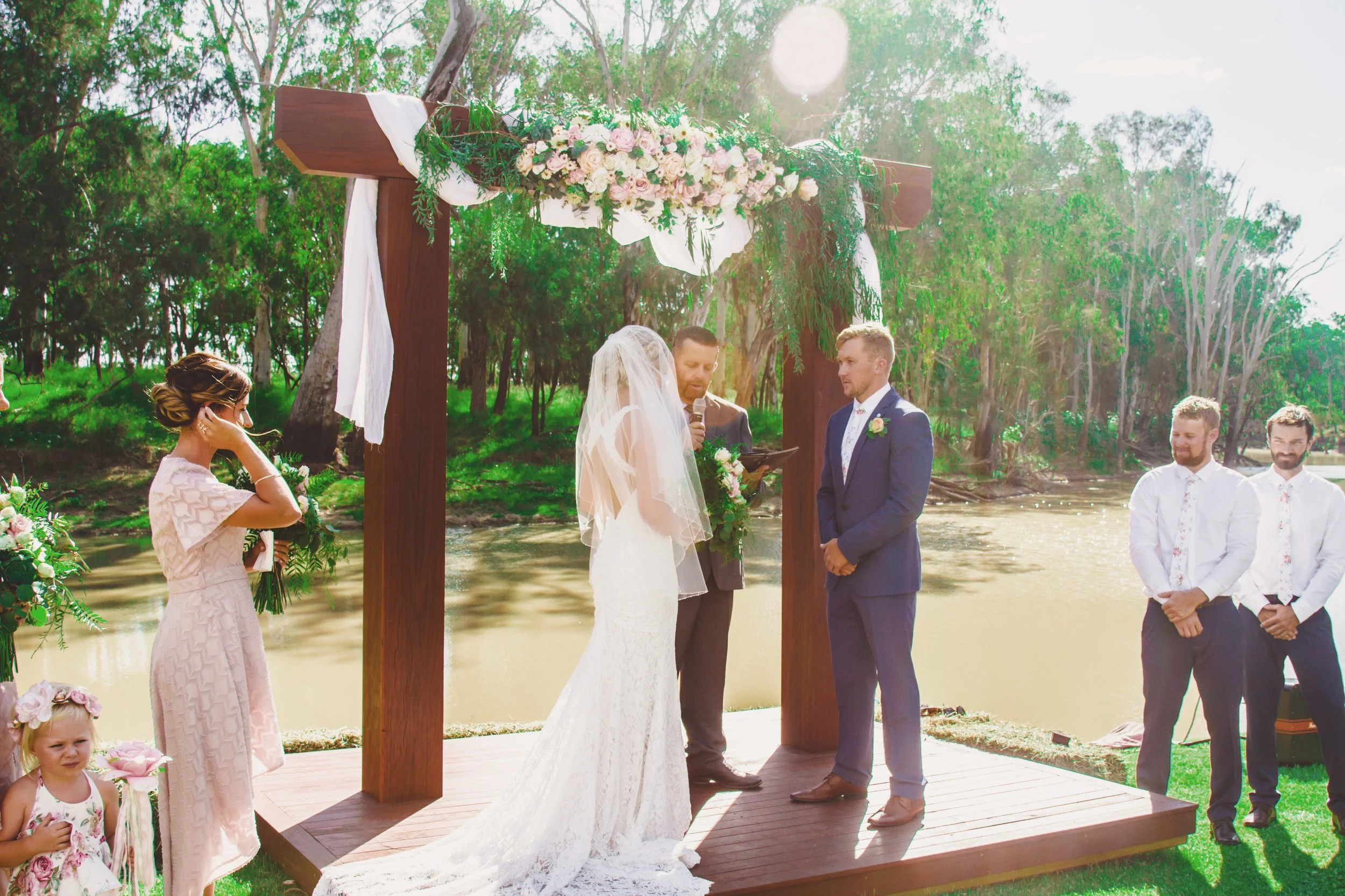 A couple gets married outdoors on a wooden platform decorated with flowers, with an officiant and a bridesmaid present, by a river and surrounded by trees.