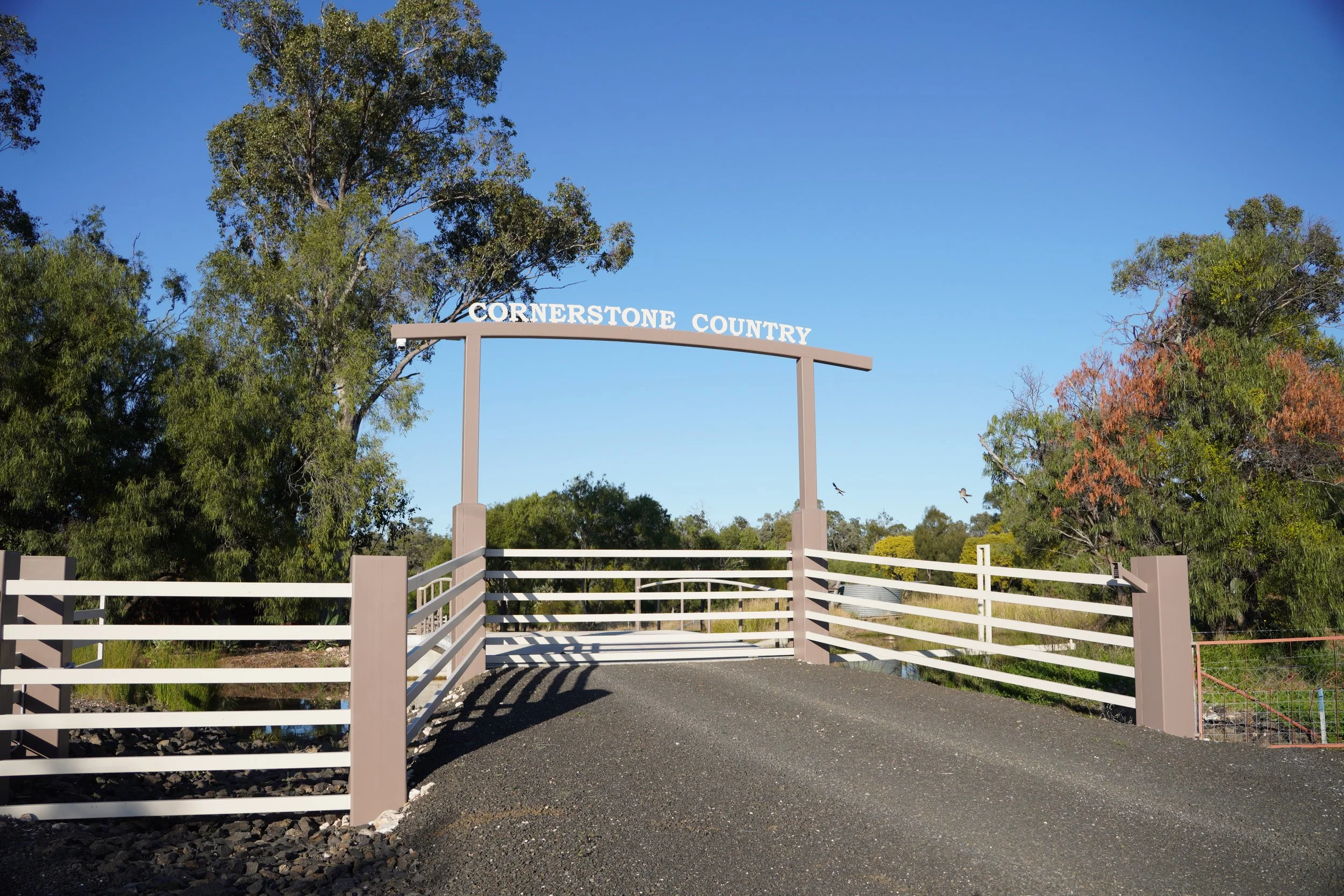 Entrance arch with the sign 'Cornerstone Country' over a white fence, surrounded by trees with green leaves and a clear blue sky.