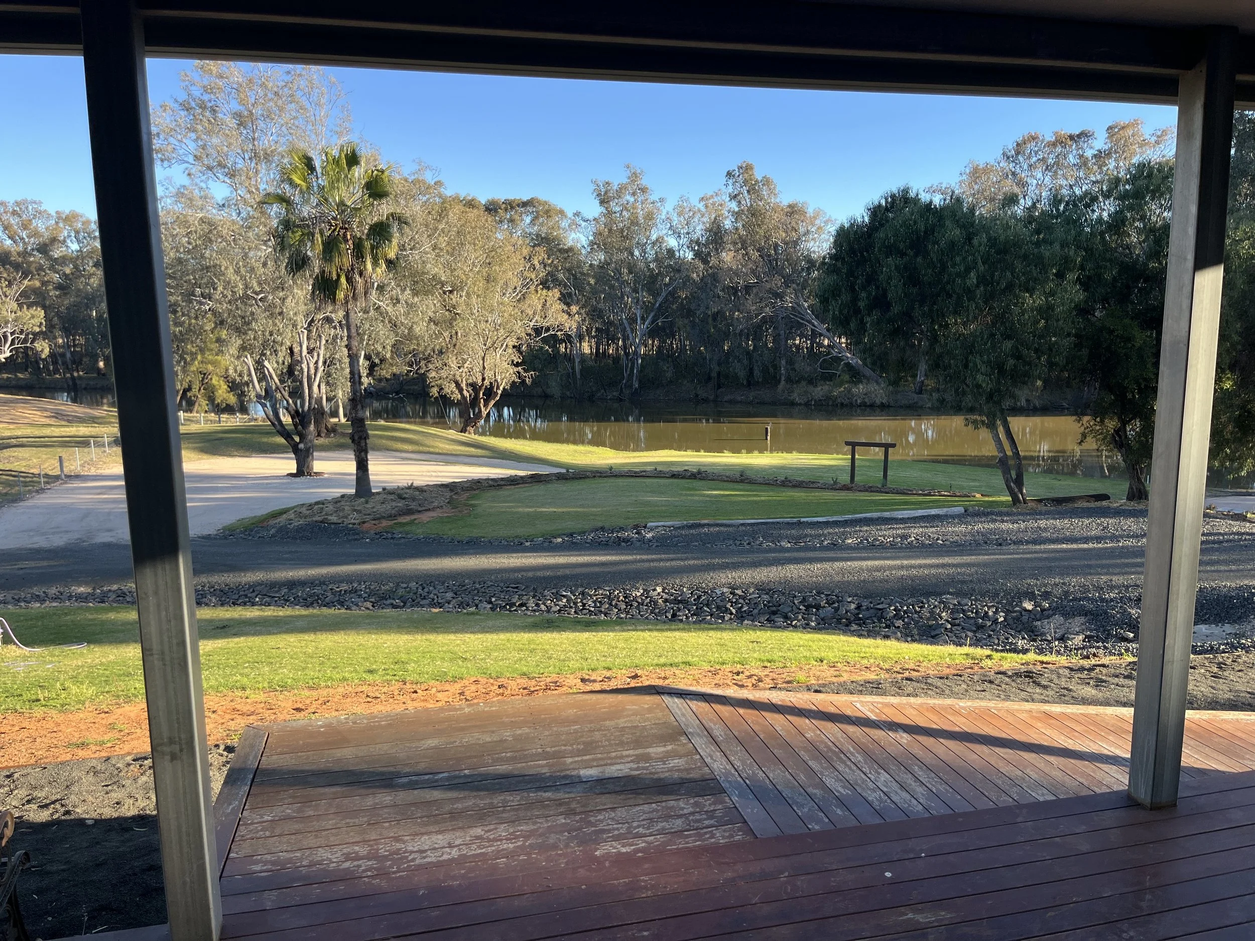 View from underneath a pavilion overlooking a park with trees, water, grass, and a walking path in the distance, with a wooden deck and metal supports in the foreground.