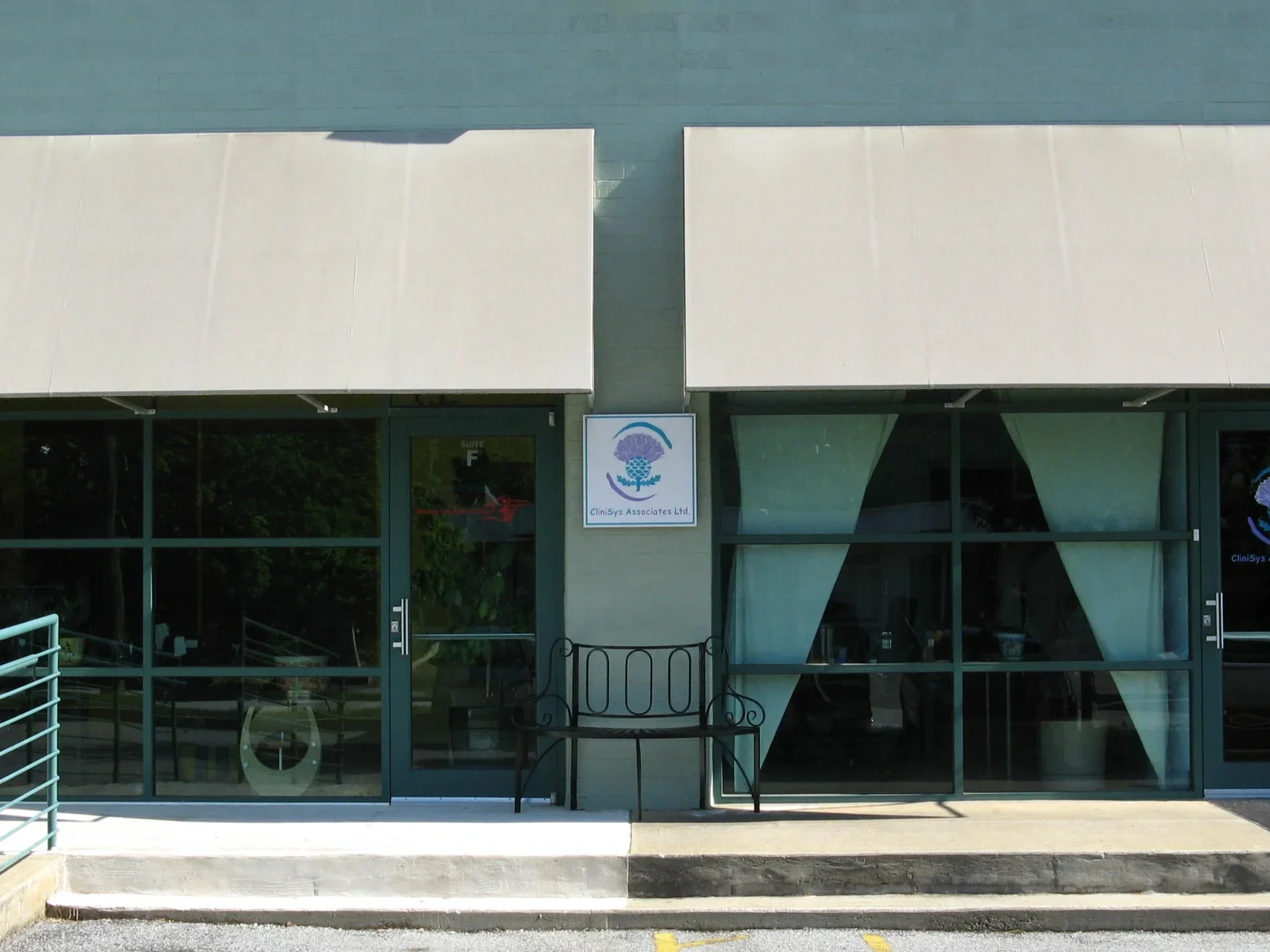 Front view of a building with large glass windows and entrance doors, a sign with a purple and blue floral logo reading 'CliniSys Associates Ltd.' on the exterior wall, and a wheelchair accessible ramp to the left.