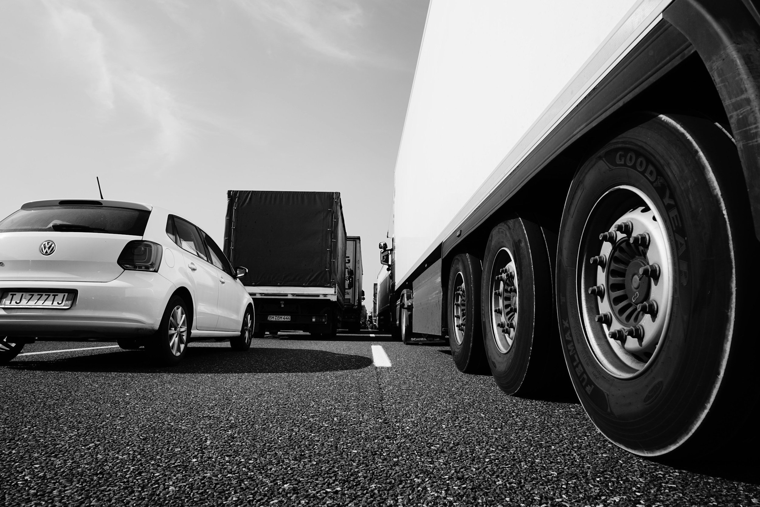 Black and white photo of vehicles on a road, featuring a white Volkswagen parked beside a large truck with visible wheel details.