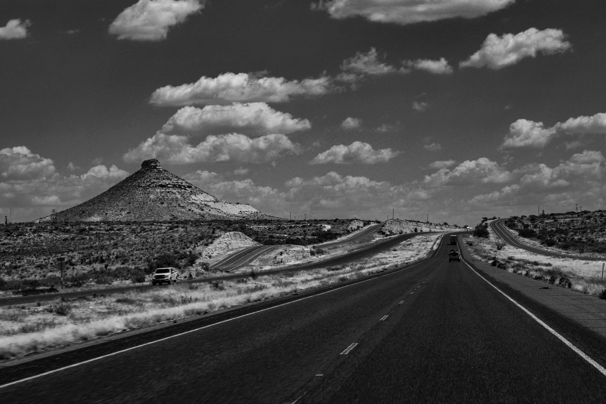 Black and white photo of a winding highway in a desert landscape with a hill and clouds in the sky.