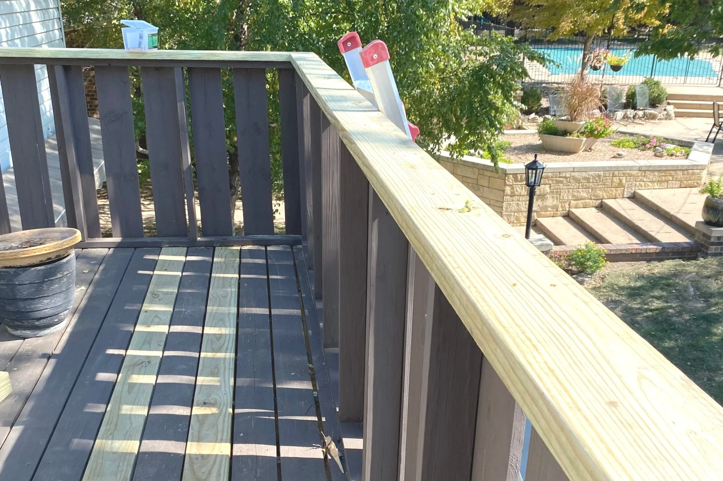 View from a wooden deck with a potted plant, overlooking a backyard with a swimming pool, garden, patio furniture, and entry stairs.