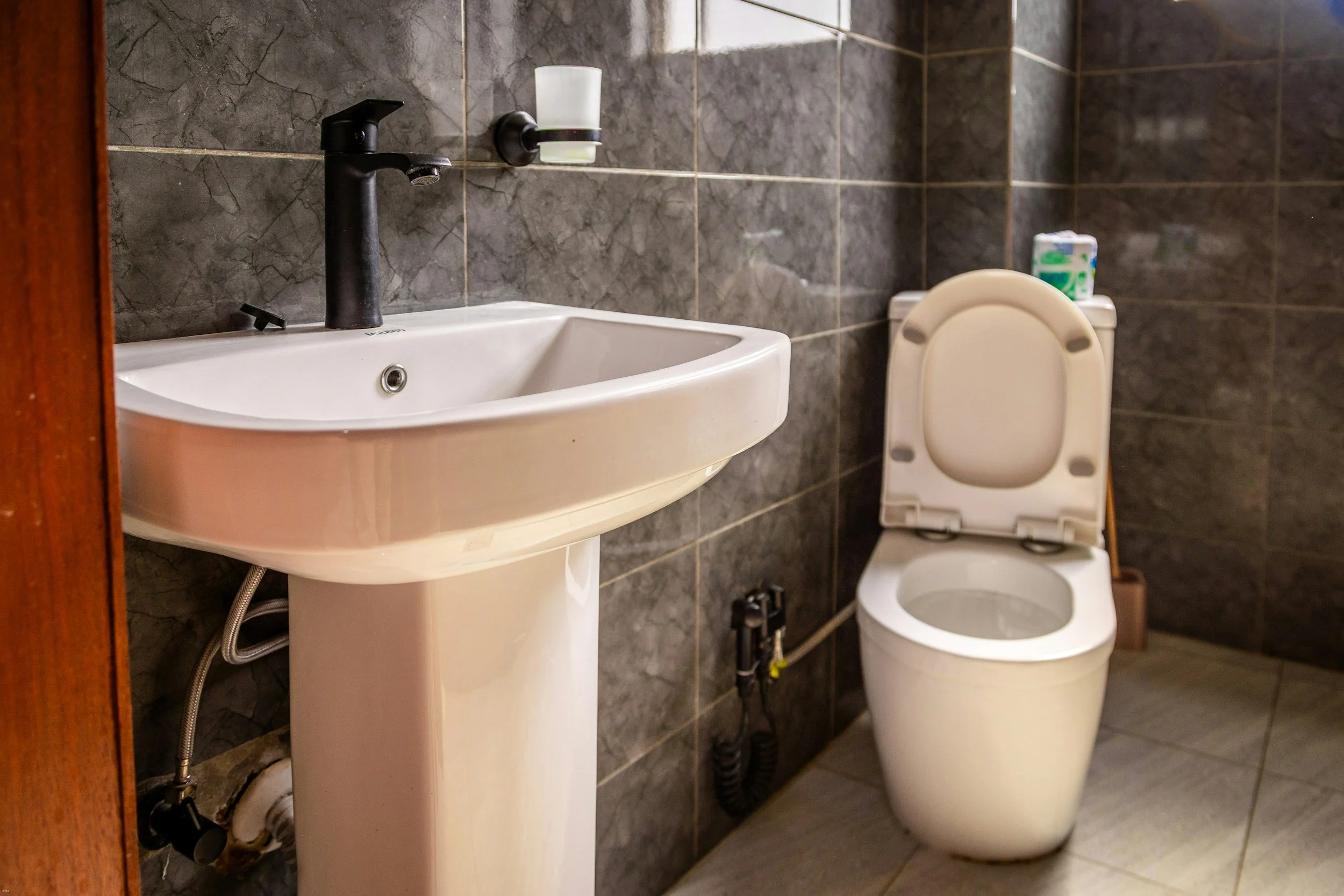 A small bathroom featuring a wall-mounted sink with a black faucet, a glass tumbler holder with a glass, and a toilet with a box of tissues on top of the tank, all against dark marble tiled walls.
