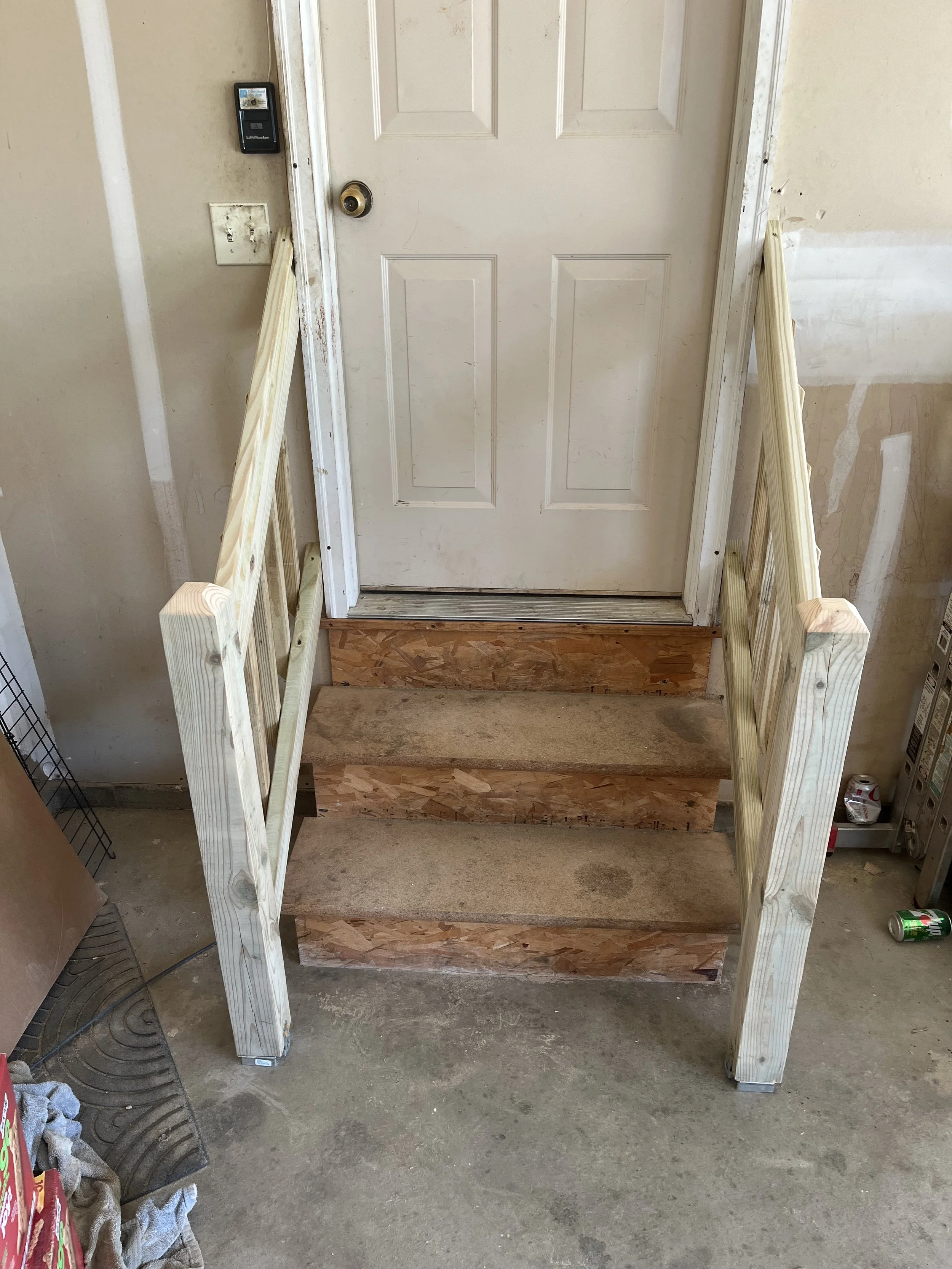 A newly constructed wooden staircase with two steps, leading to a white door, with handrails on either side, inside a building under renovation.