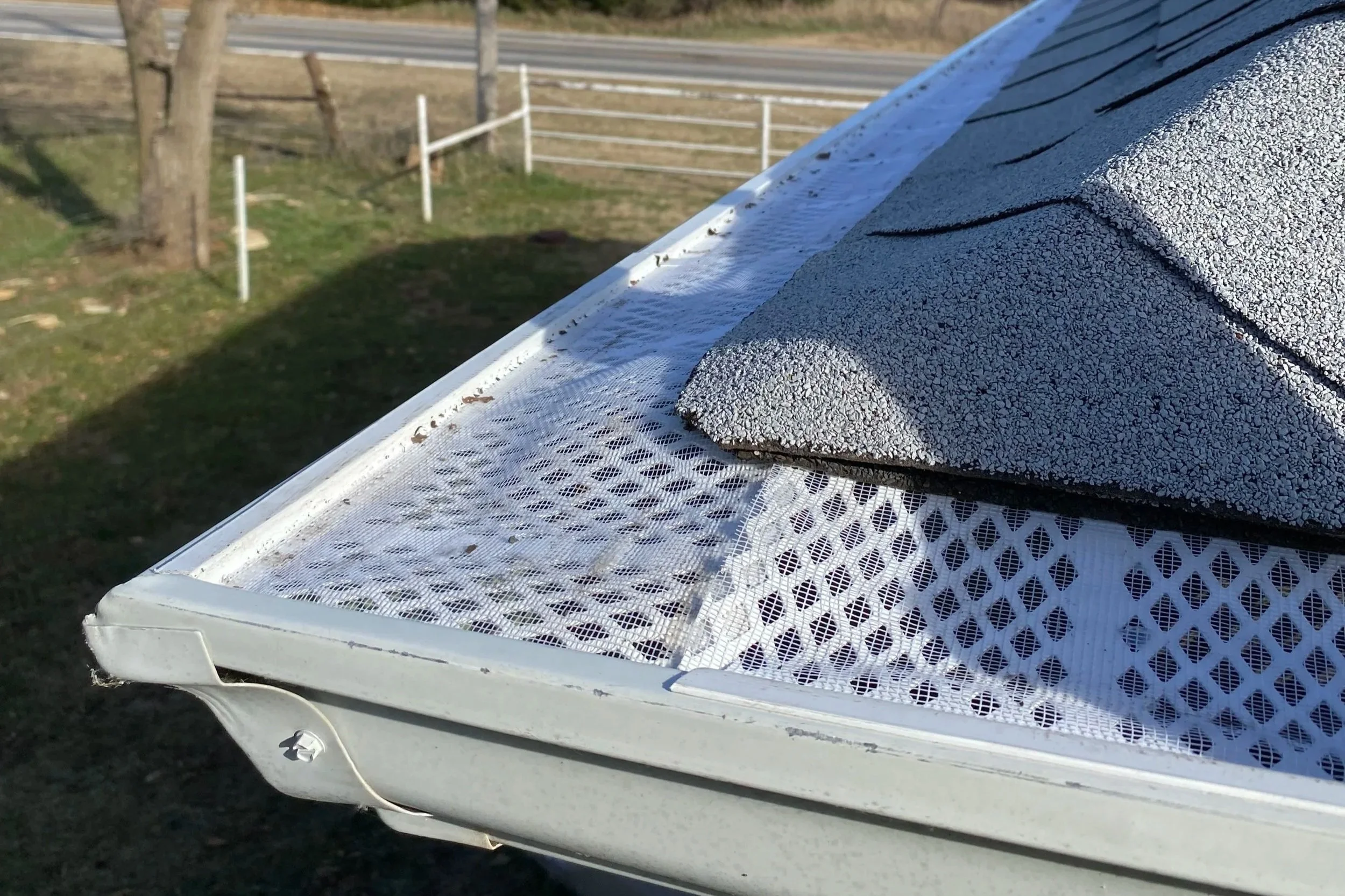 Close-up of a house's roof with gravel shingles and a white metal mesh soffit, next to a grassy yard with trees and a road in the background.