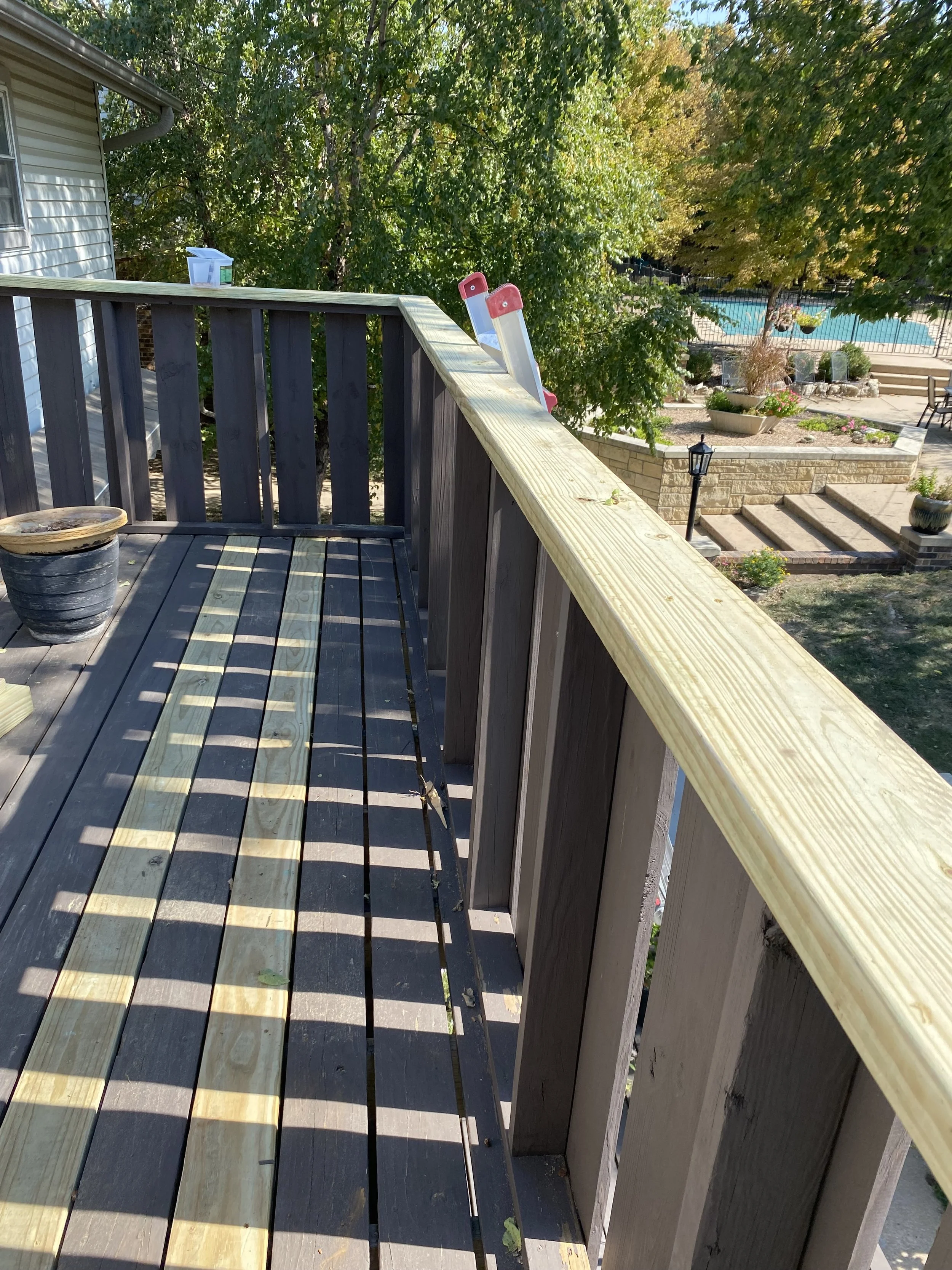 A rooftop deck with a potted plant, a plastic container, and a ladder hanging over the edge, overlooking a backyard with a swimming pool, garden beds, and trees in fall foliage.