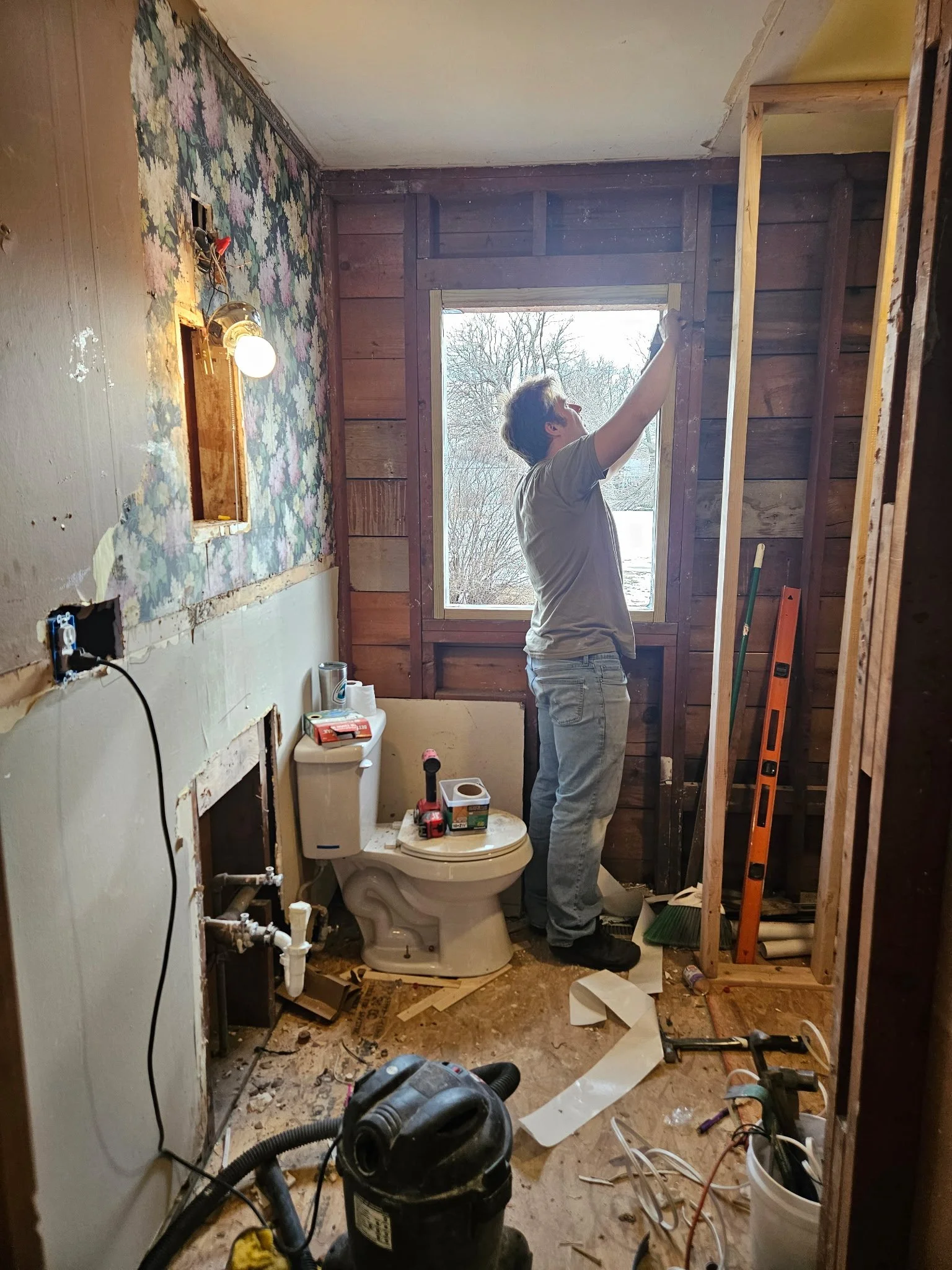 A person is working on renovation or construction inside a bathroom, removing part of the wall. The bathroom has exposed wood framing and walls, a toilet, and construction tools and clutter on the floor.