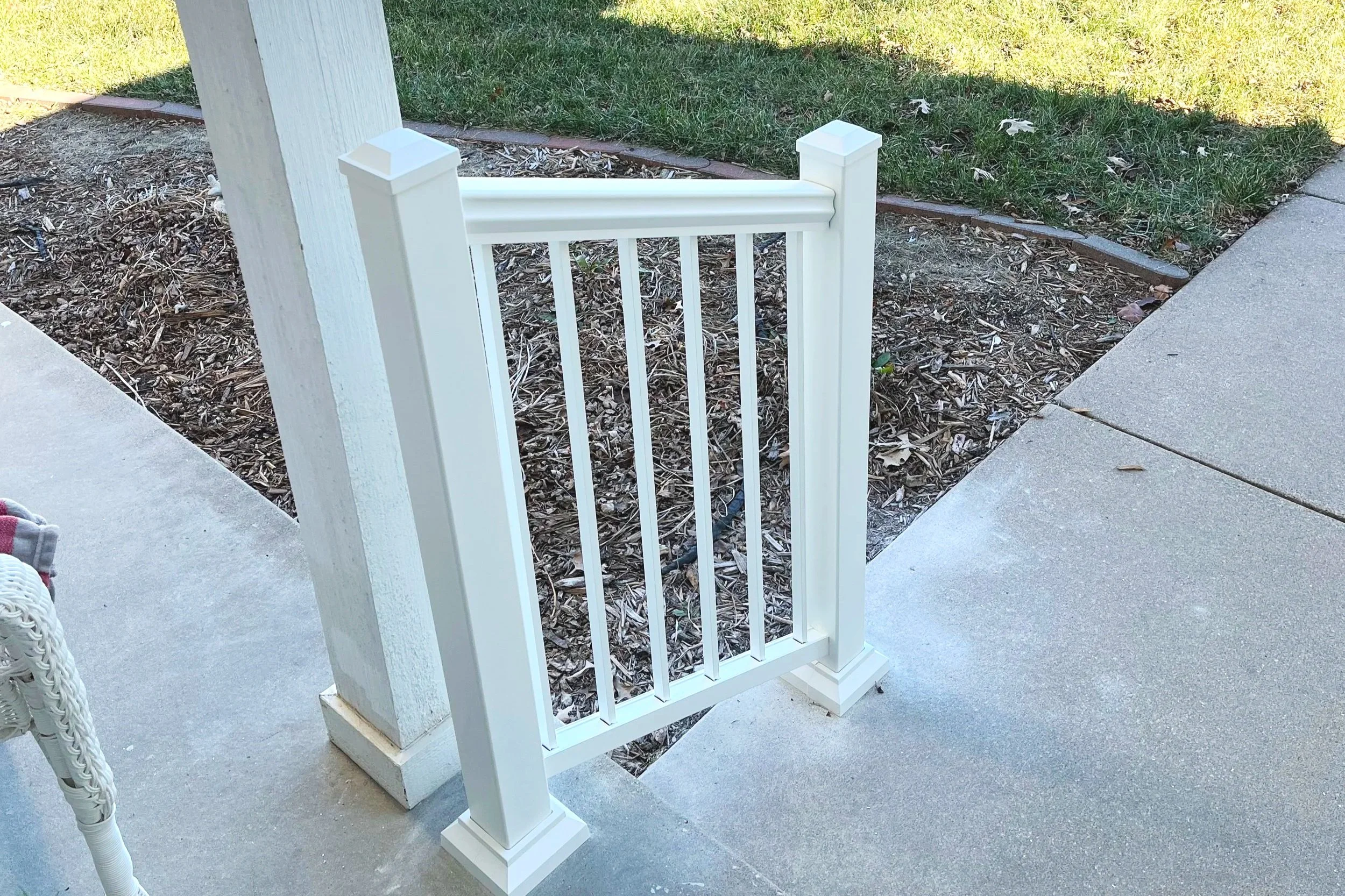 A white picket gate is attached to a white fence post on a concrete porch. There is a yard with grass, mulch, and a garden bed in the background.