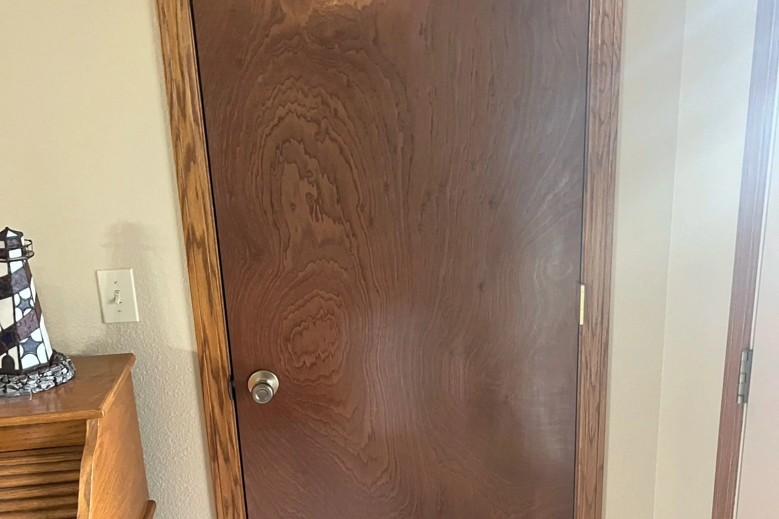 A wooden door with a silver doorknob in a room with beige walls, a wooden desk to the left, and a decorative lamp on the desk.