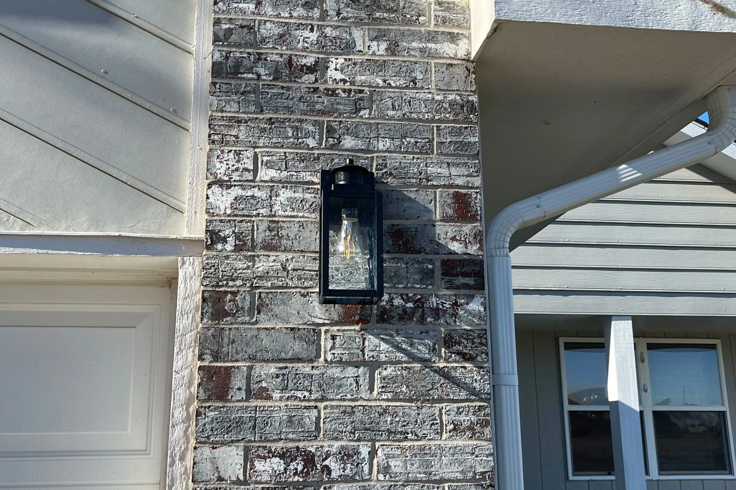 Exterior house wall with a brick and siding combination, a black outdoor lantern, white PVC gutter pipe, and a window with white trim.