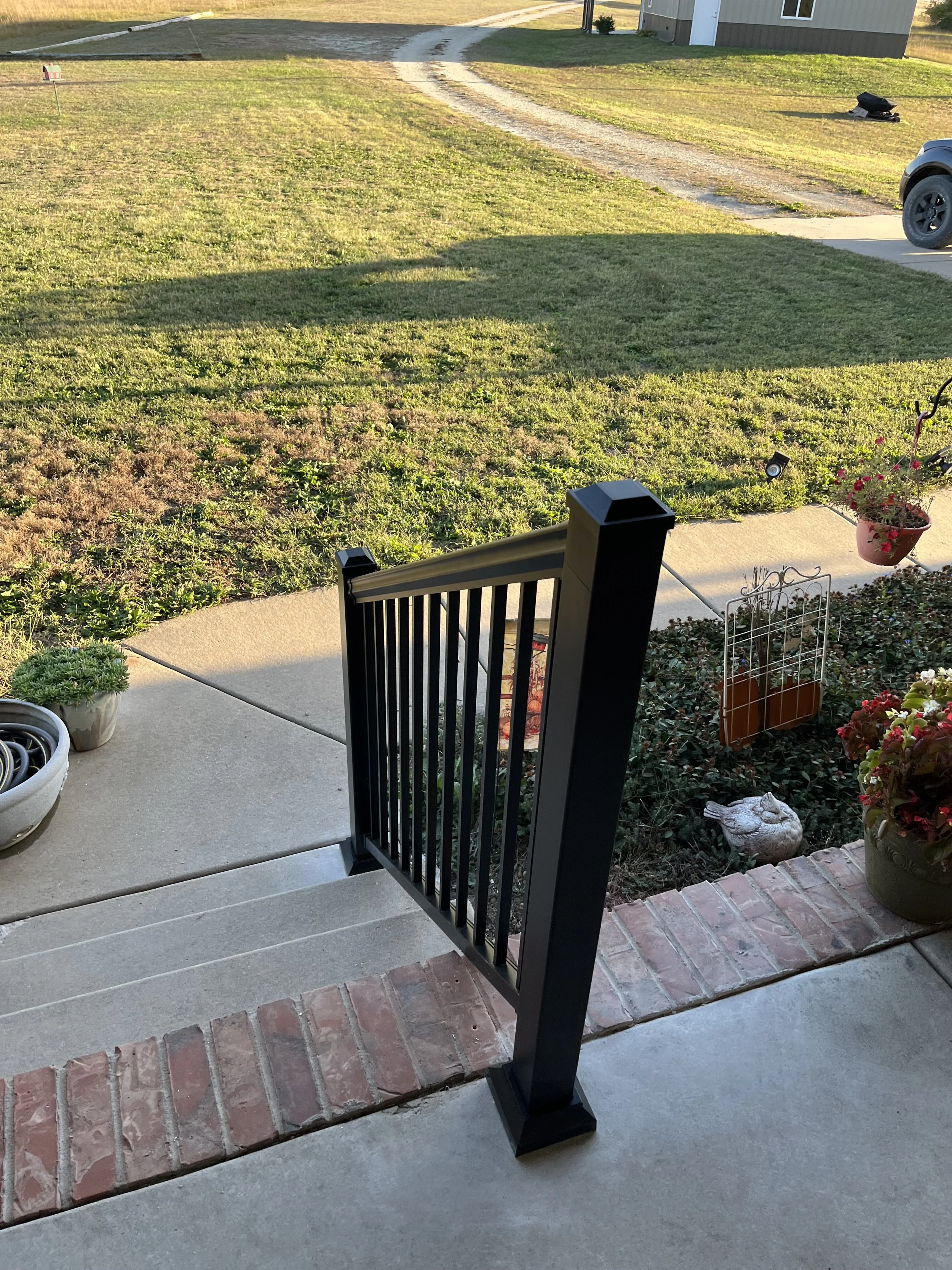 View from a porch looking out onto a yard and driveway. There are steps leading down from the porch, with potted plants and garden decorations nearby. The yard is grassy with patches of green and brown. A gravel driveway curves into the distance, with a house visible in the background and a trash bin on the side.