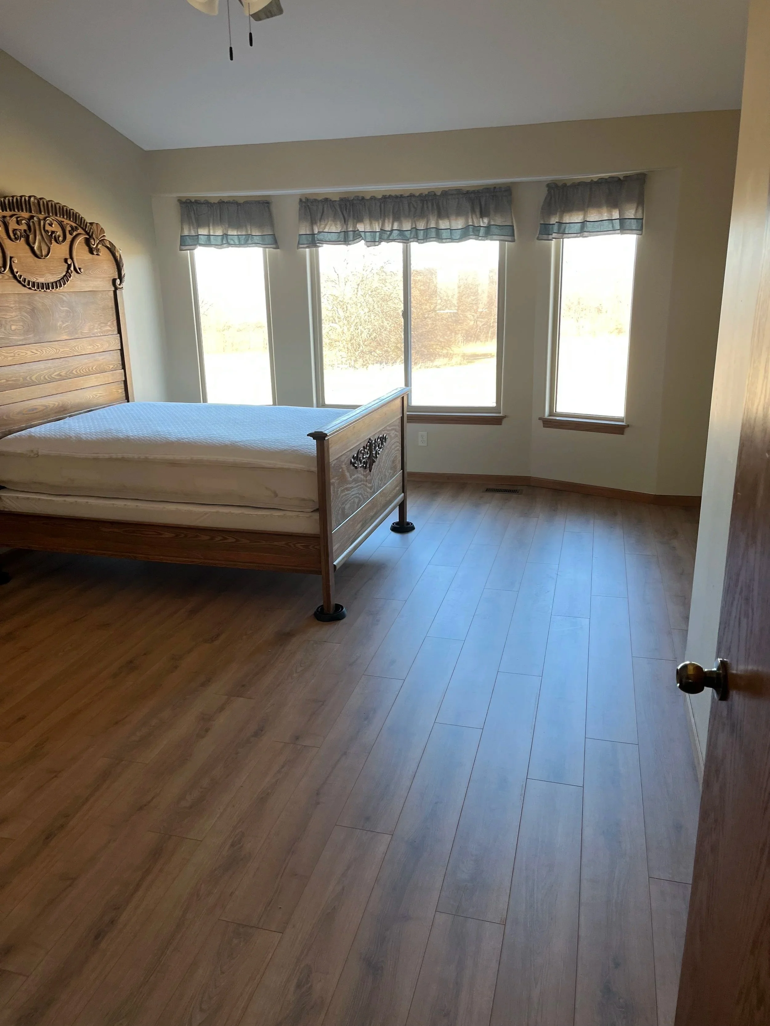 Empty bedroom with wooden bed frame, mattress, three large windows with valances, hardwood floors, ceiling fan, and sunlight streaming in.