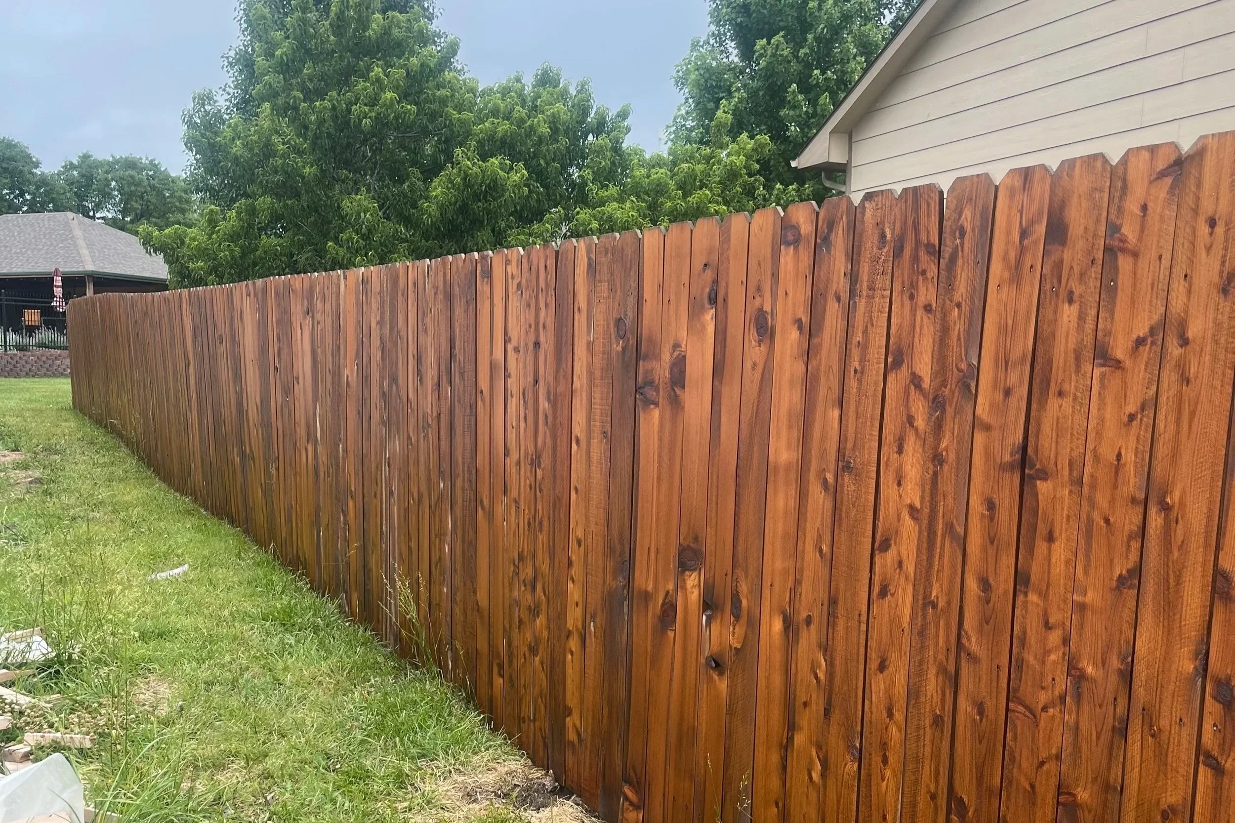 A newly stained tall wooden privacy fence with vertical planks along a grassy yard, with trees and a house with light beige siding in the background under cloudy skies.
