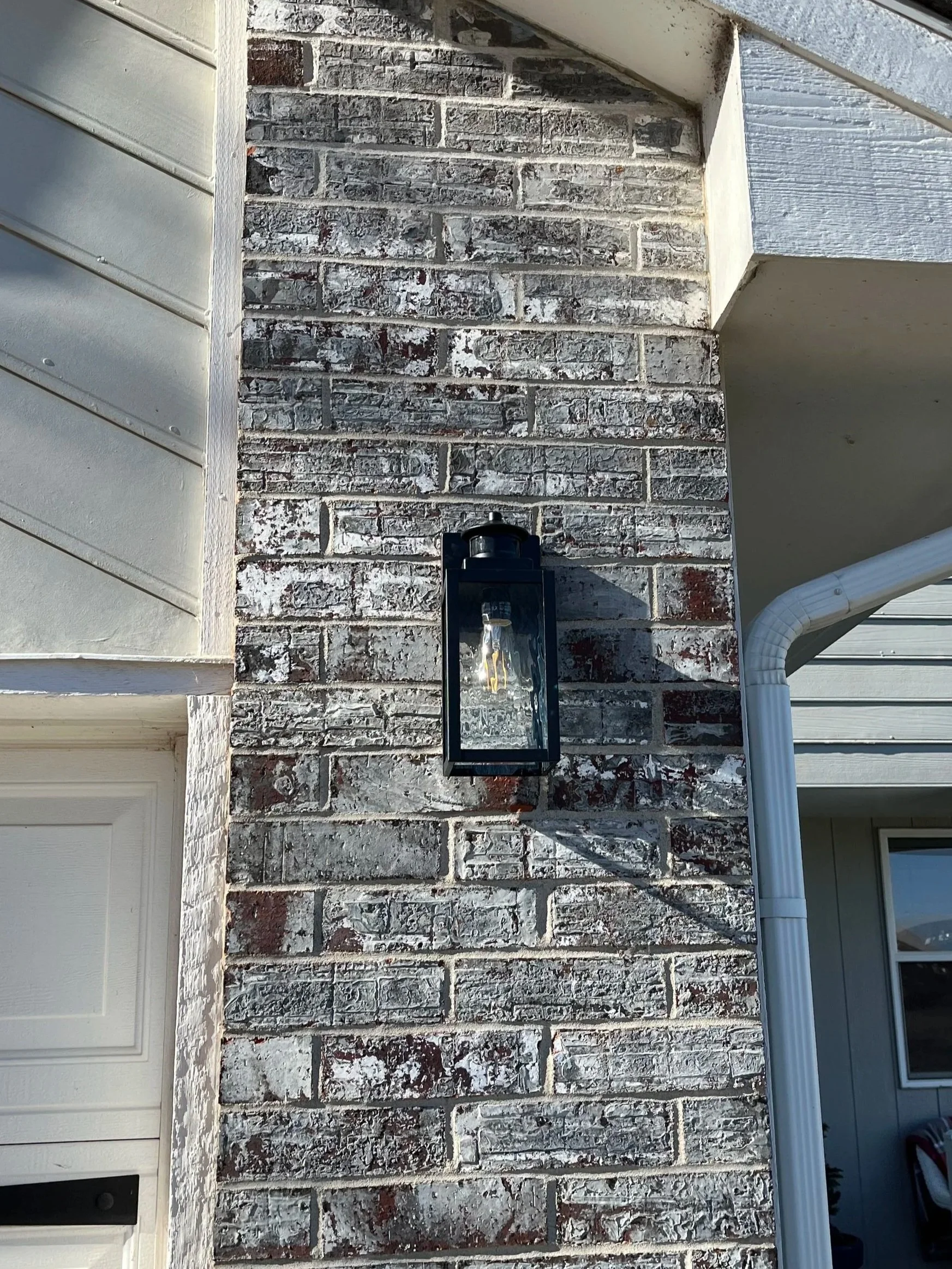 Close-up of a brick exterior wall with a black outdoor lantern mounted on it, next to a white gutter pipe, and a window with a white frame on the right.