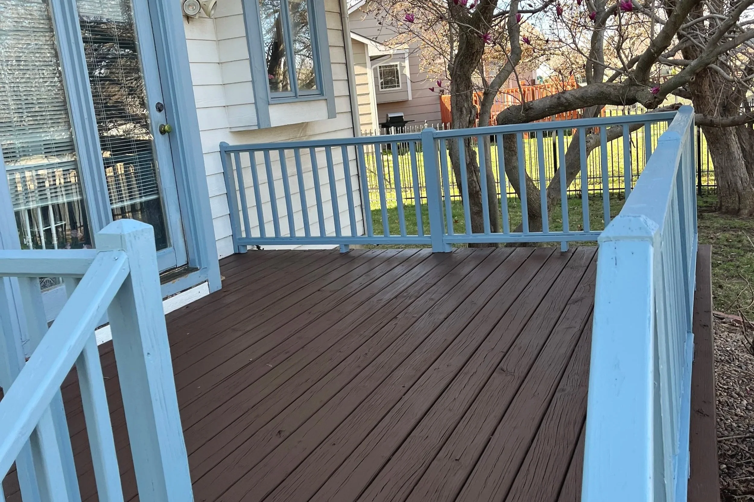 A wooden deck with a blue railing attached to the side of a house. A tree with pink flowers and a large branch extending across the yard. In the background, neighboring houses and a black grille are visible.