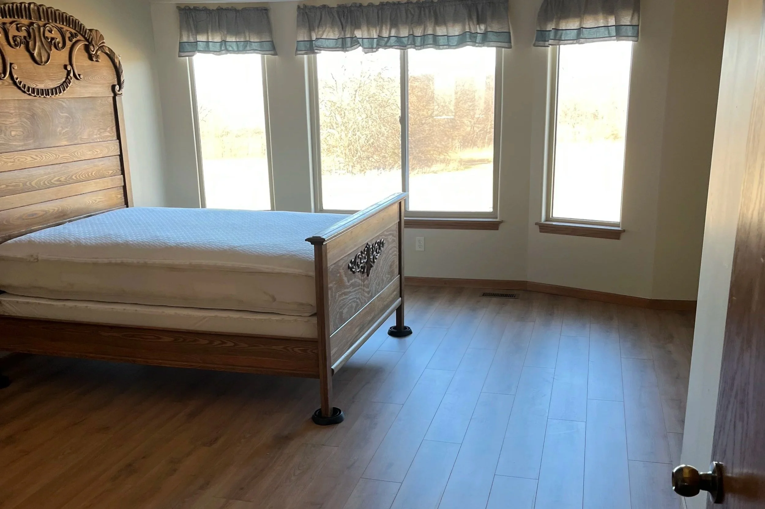 A bedroom with a large window covered by valance curtains, a wooden bed frame with a mattress, and hardwood floors.