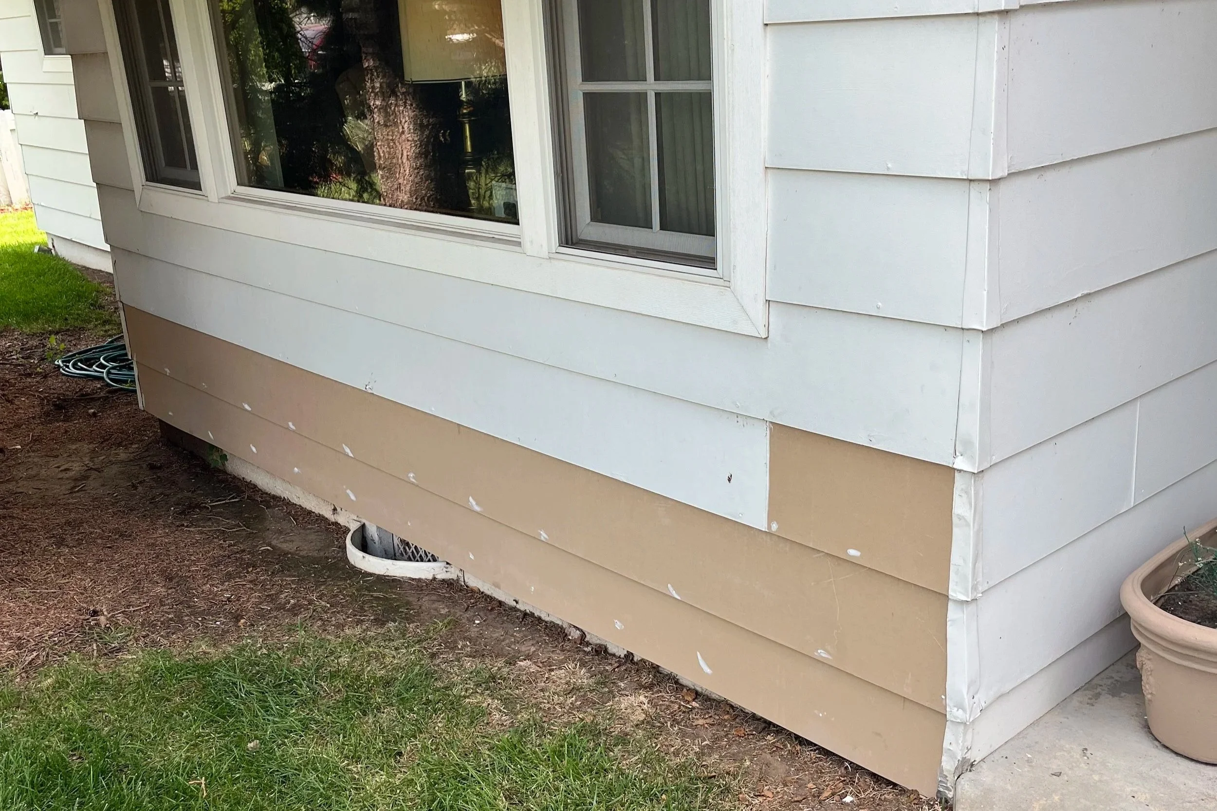 The image shows the side of a house with siding that has been painted or is in the process of being painted. There is a window with a screen and reflections of trees, and a hose coiled on the ground nearby. The house is elevated with space underneath, and a potted plant is visible on the right side.