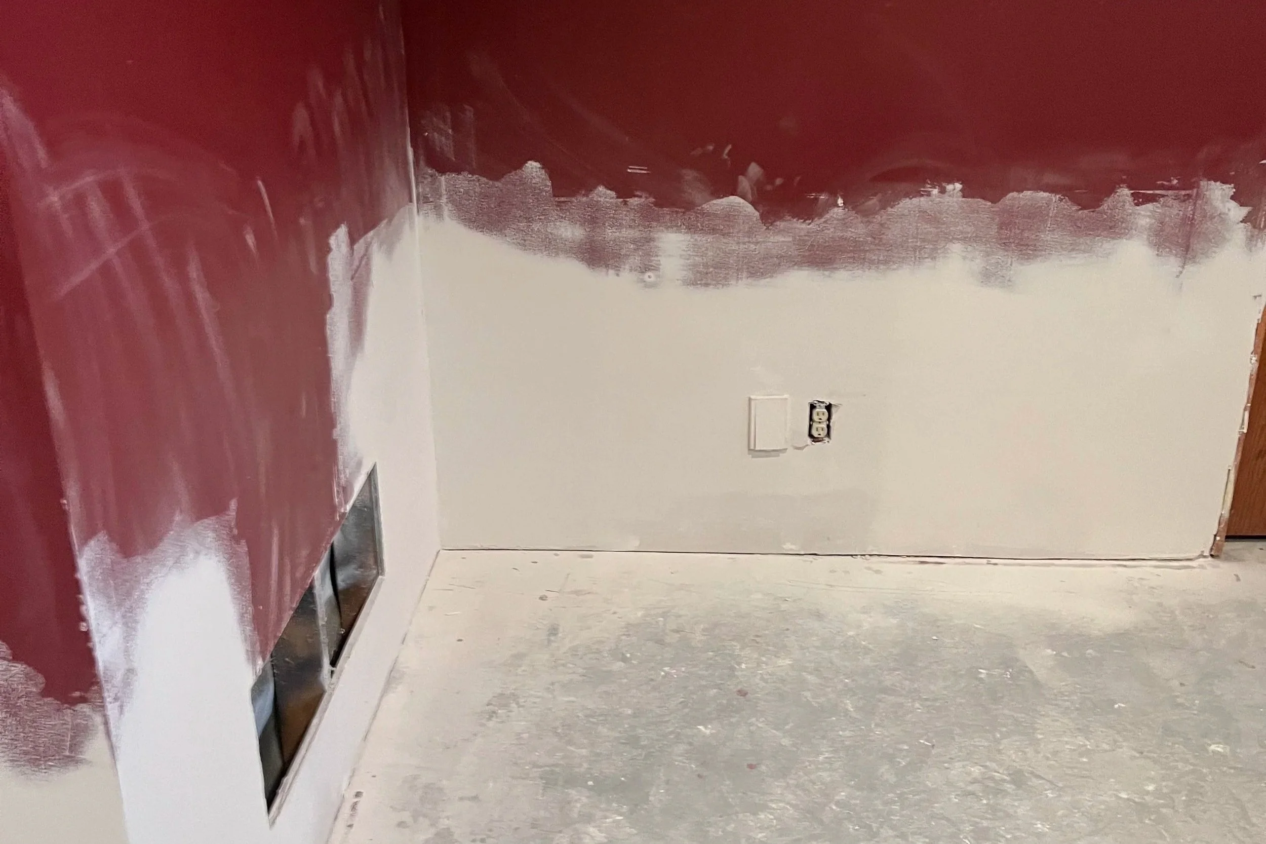 Interior room with partially painted wall, white paint on wall, red paint on ceiling and part of wall, floor covered with drop cloth and paint splatters, electrical outlet on wall.
