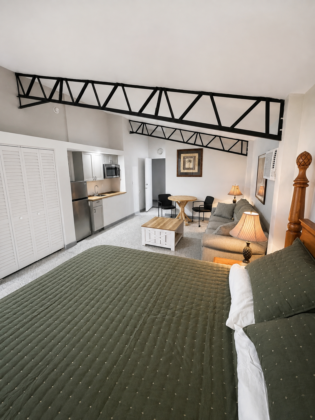 Interior of a suite with a bed in the foreground, small kitchenette with stainless steel appliances, sitting area with a sofa, table, and chairs, and decorative lamps.