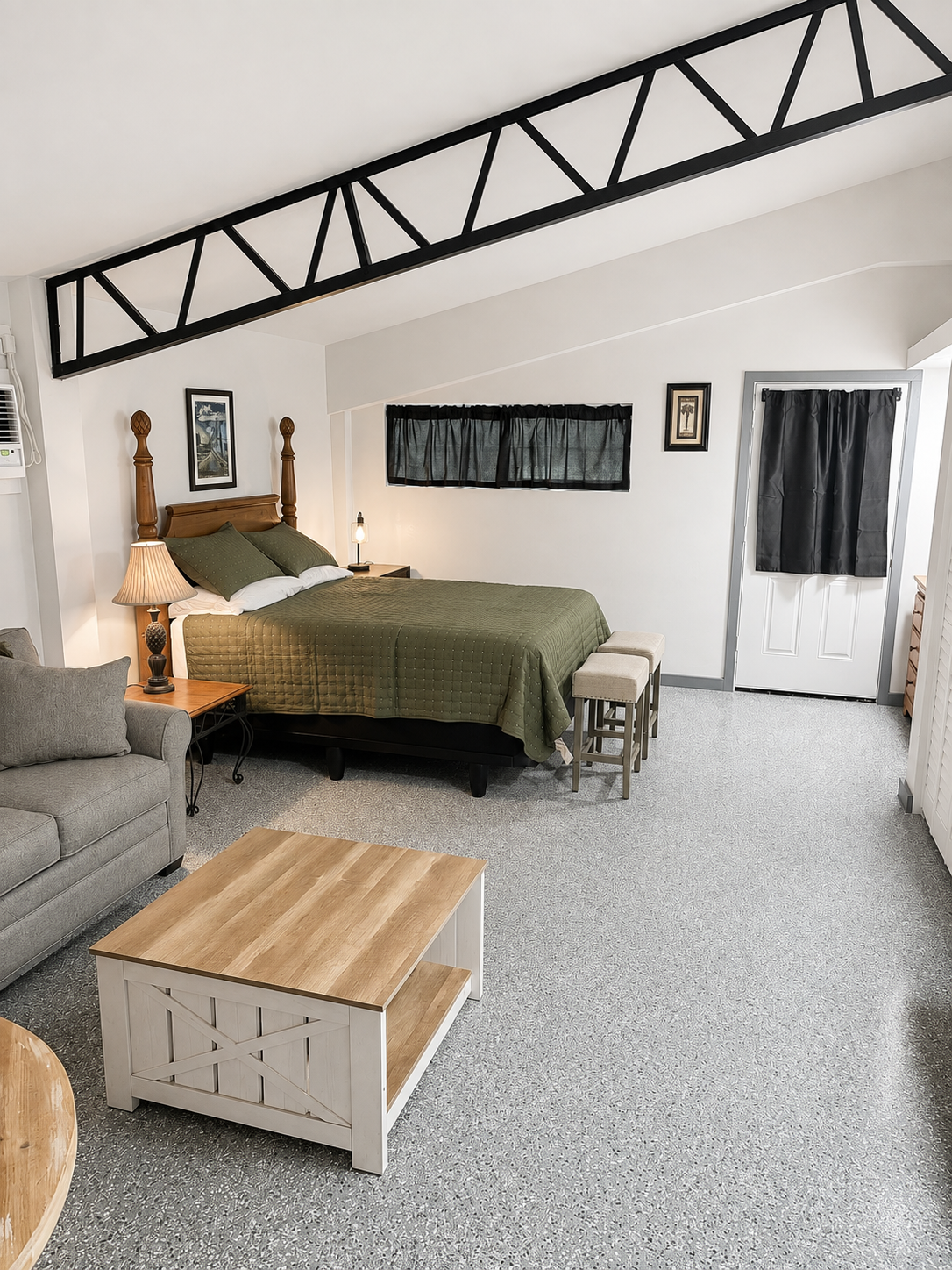 A cozy suite with a high vaulted ceiling. There is a wooden bed with green bedding and pillows, a bedside table with a lamp, a couch with pillows, a coffee table, and a window with a black curtain. The room has white walls and carpeted flooring.