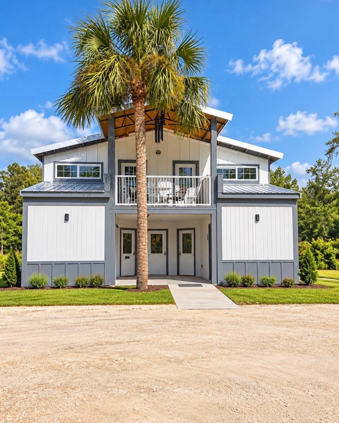 Modern two-story house with white and gray siding, a balcony with chairs, and a large palm tree in front, set against a blue sky with scattered clouds.
