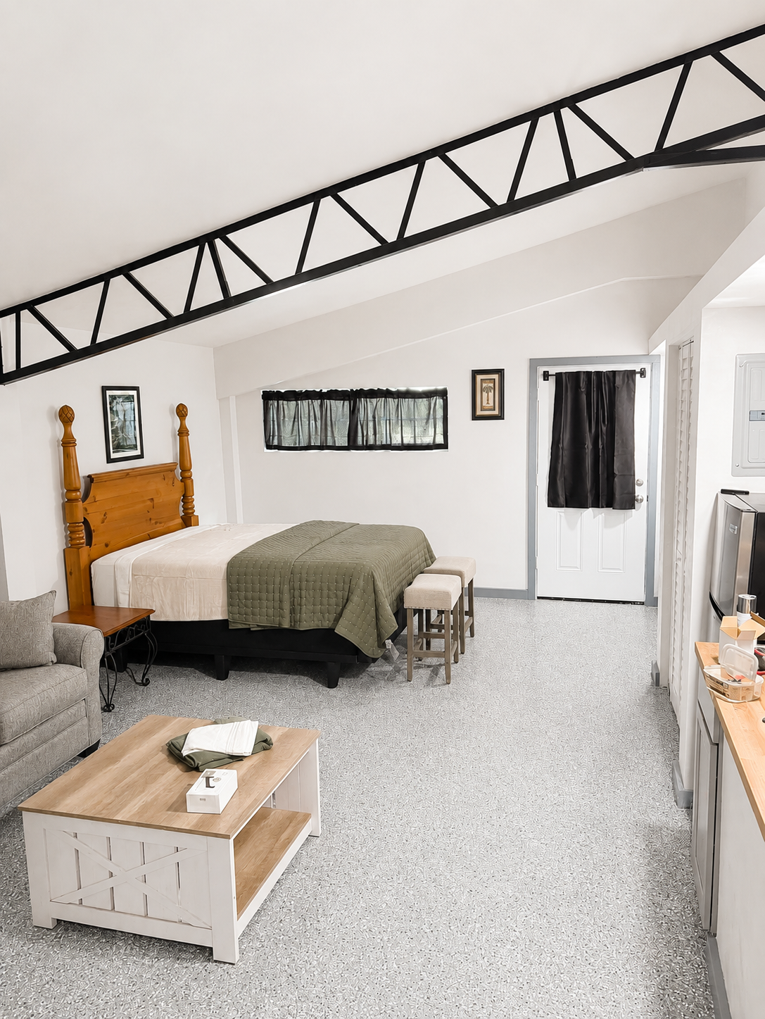 A suite with a headboard and quilted blanket. Stools are against the wall near the door. A couch and a coffee table with tissues and a magazine are in the foreground. The room has gray carpeting and a sloped ceiling with black supporting beams.