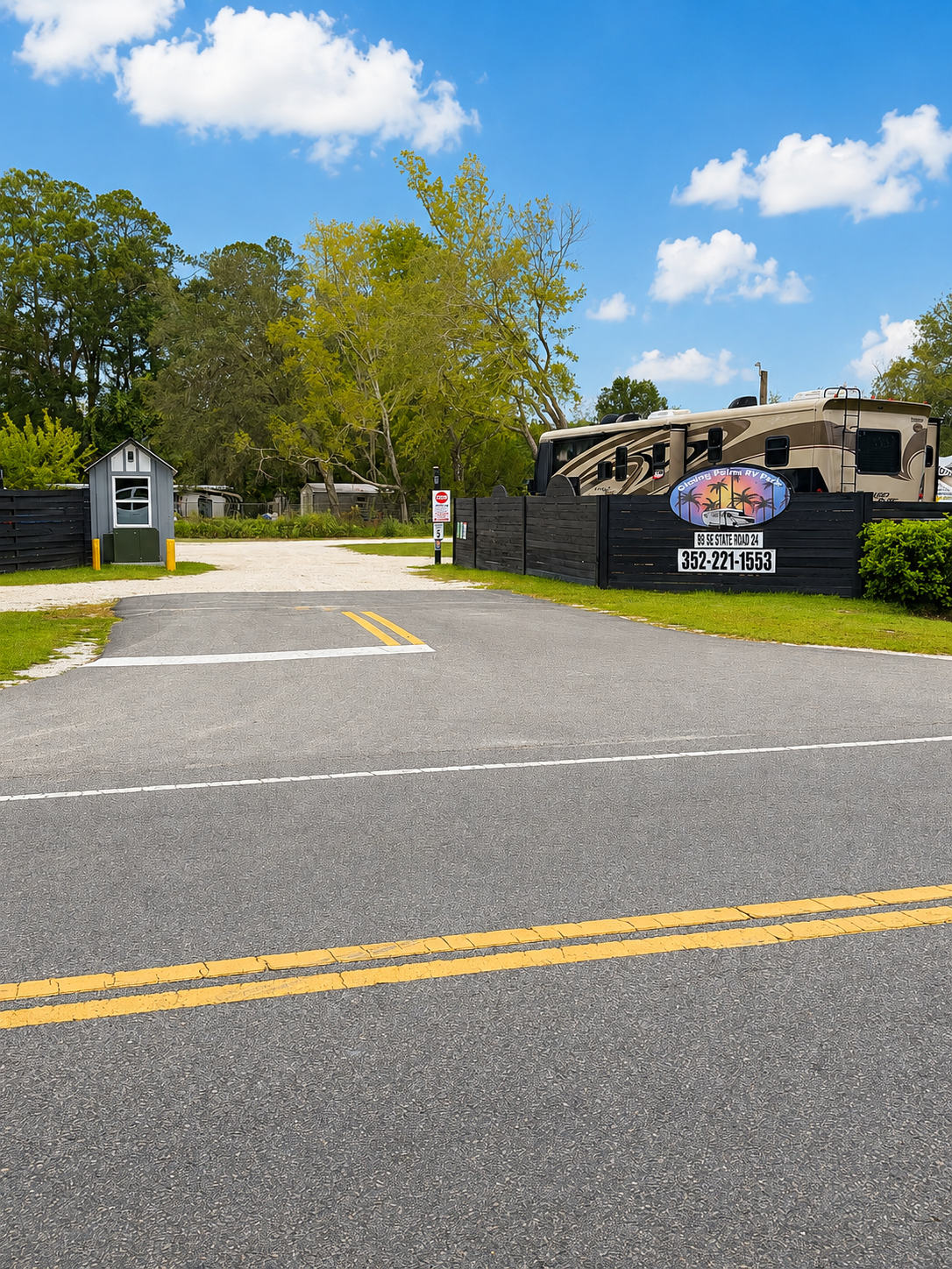 A large RV parked inside a black fenced area at a campsite with trees and a partly cloudy sky in the background.