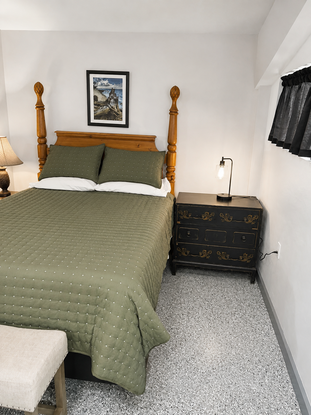 A suite with a wooden bed frame, green quilted bedding, matching pillows, a beige stool at the foot of the bed, a black nightstand with gold accents, a lamp, a small bedside lamp, and a framed landscape picture on the wall.