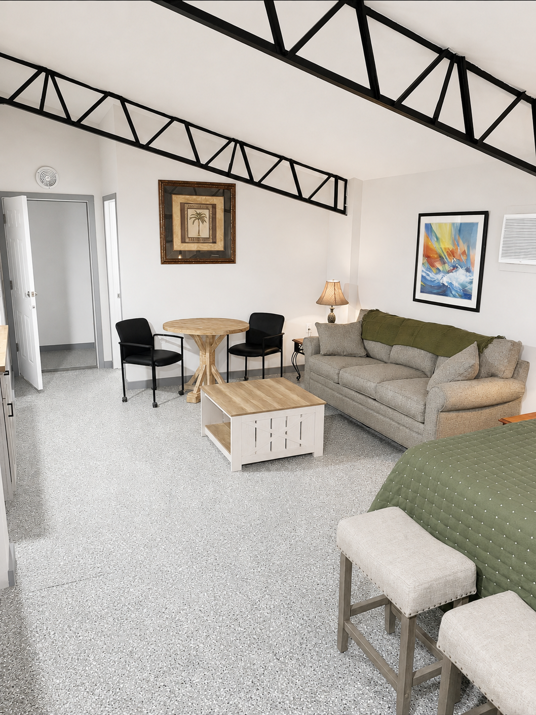  A suite with a beige sofa, blanket, pillows, a wooden coffee table, colorful abstract art, a table lamp, a small round dining table with two black chairs, and a patterned carpet. There are framed pictures on white walls and black exposed beams.