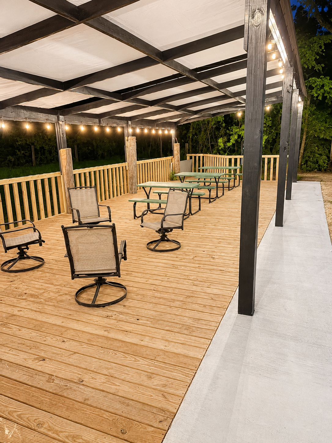 An outdoor patio covered by a black and white canopy, lit by string lights. The patio has a wooden floor, a wooden railing, and several green and beige tables with metal frames. There are also beige swivel chairs with armrests around the tables.
