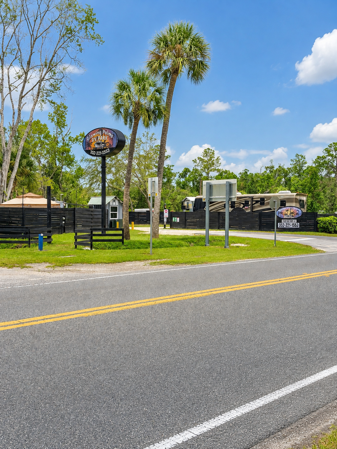 A roadside scene featuring a sign for Glowing Palms RV Park, palm trees, a large motorhome, a black fence, green grass, and a clear blue sky with scattered clouds.