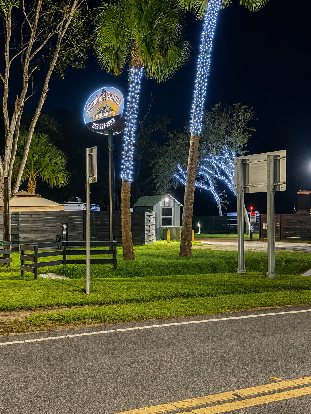 Nighttime scene of a street side with trees wrapped in white string lights, a small building, a road, and a sign for Glowing Palms.