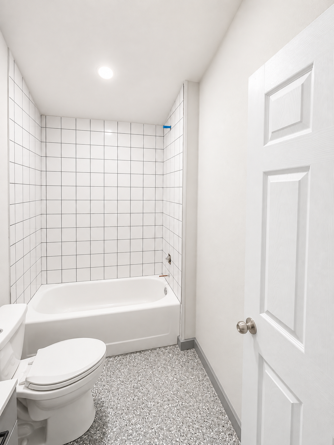 A suite bathroom featuring a toilet, bathtub with tiled walls, and a door, with terrazzo flooring, illuminated by a ceiling light.