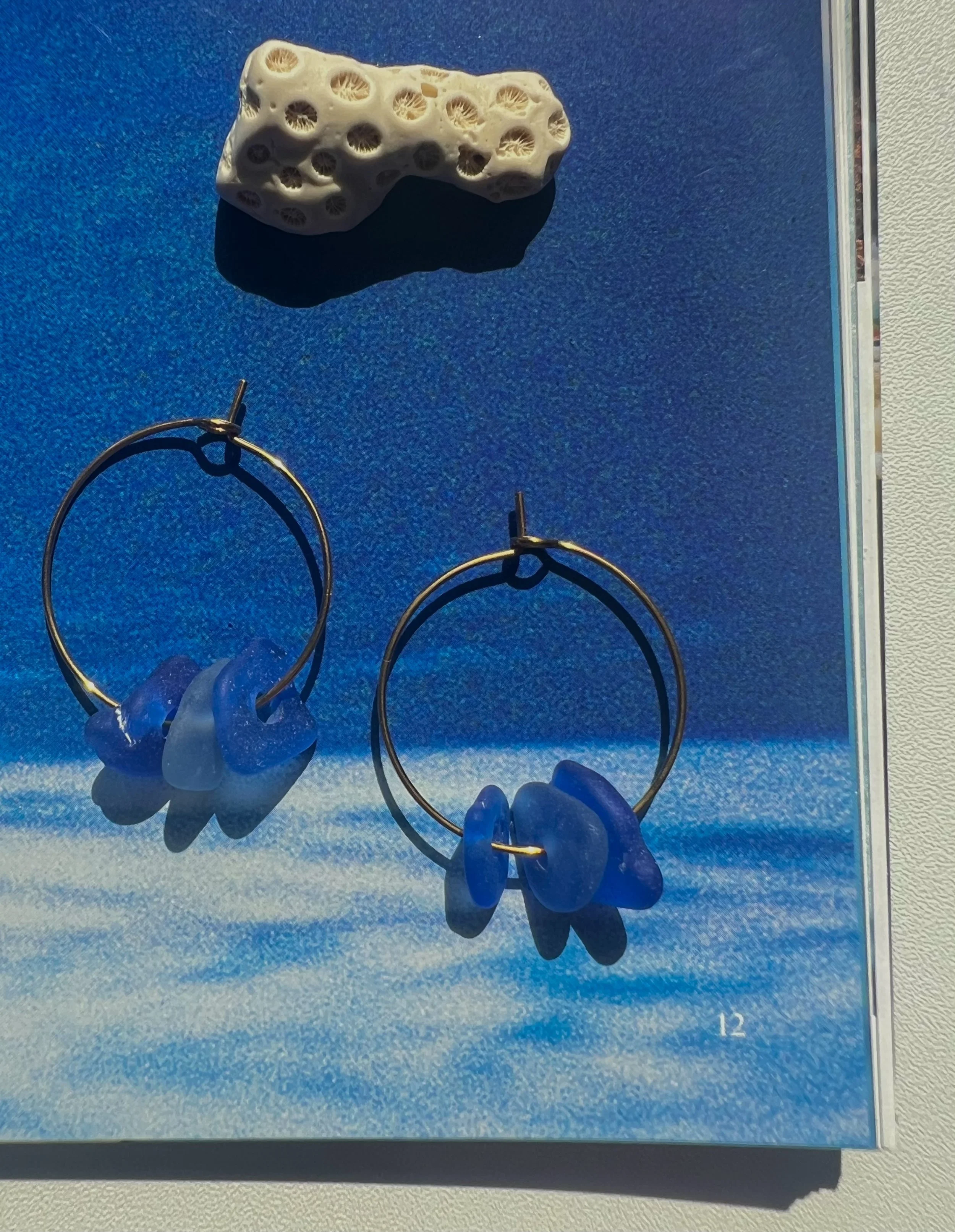 A white coral piece with multiple holes set on a blue background, accompanied by a pair of gold hoop earrings with blue hooks underneath.