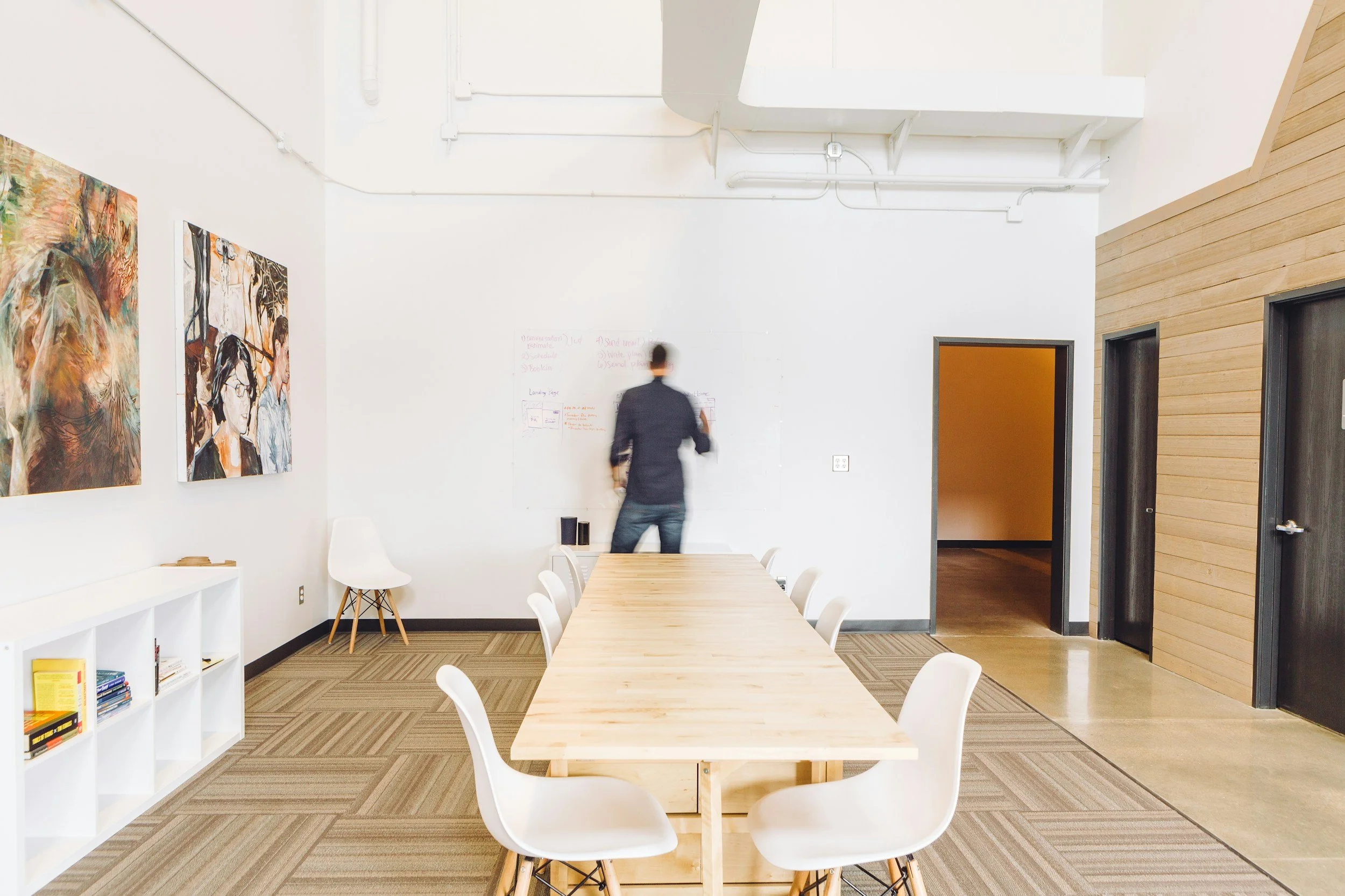 Modern conference room with white walls, wooden accents, a long wooden table surrounded by white chairs, artwork on the walls, and a person writing on a whiteboard.