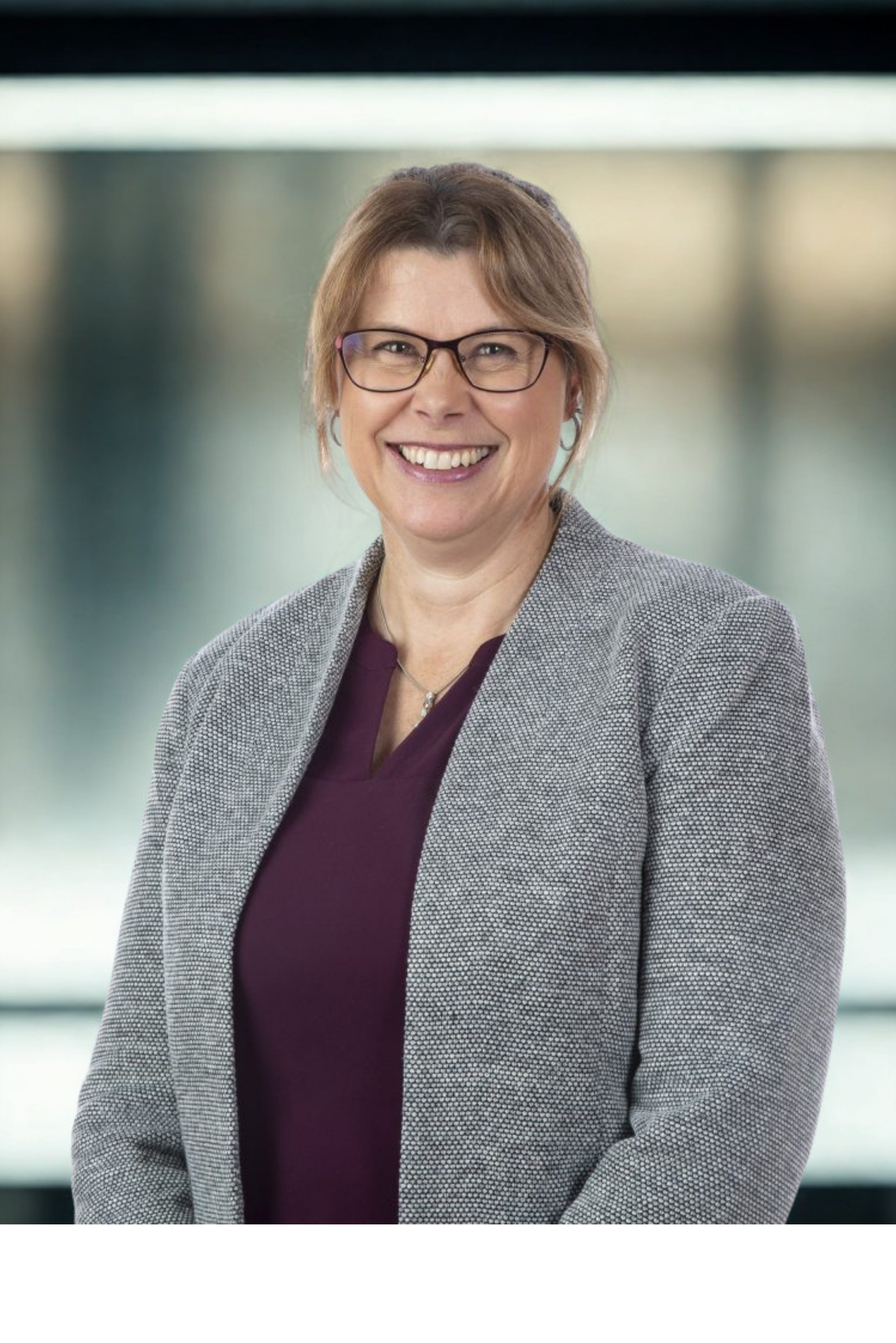 Professional woman with glasses smiling, wearing a gray blazer and maroon top, standing in a modern office environment.