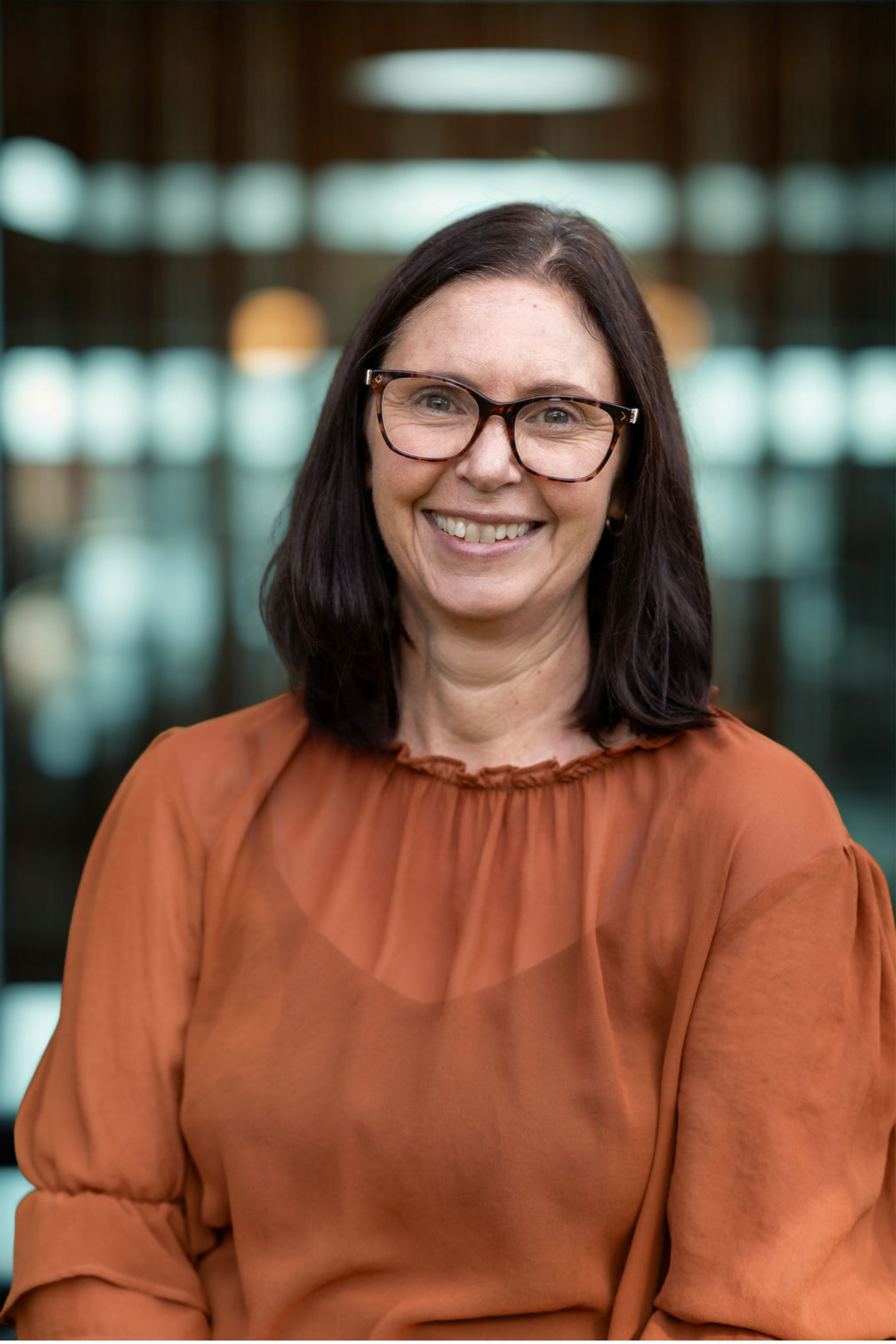 Close-up of a woman with dark hair, glasses, smiling, wearing an orange blouse, indoors with blurred background.