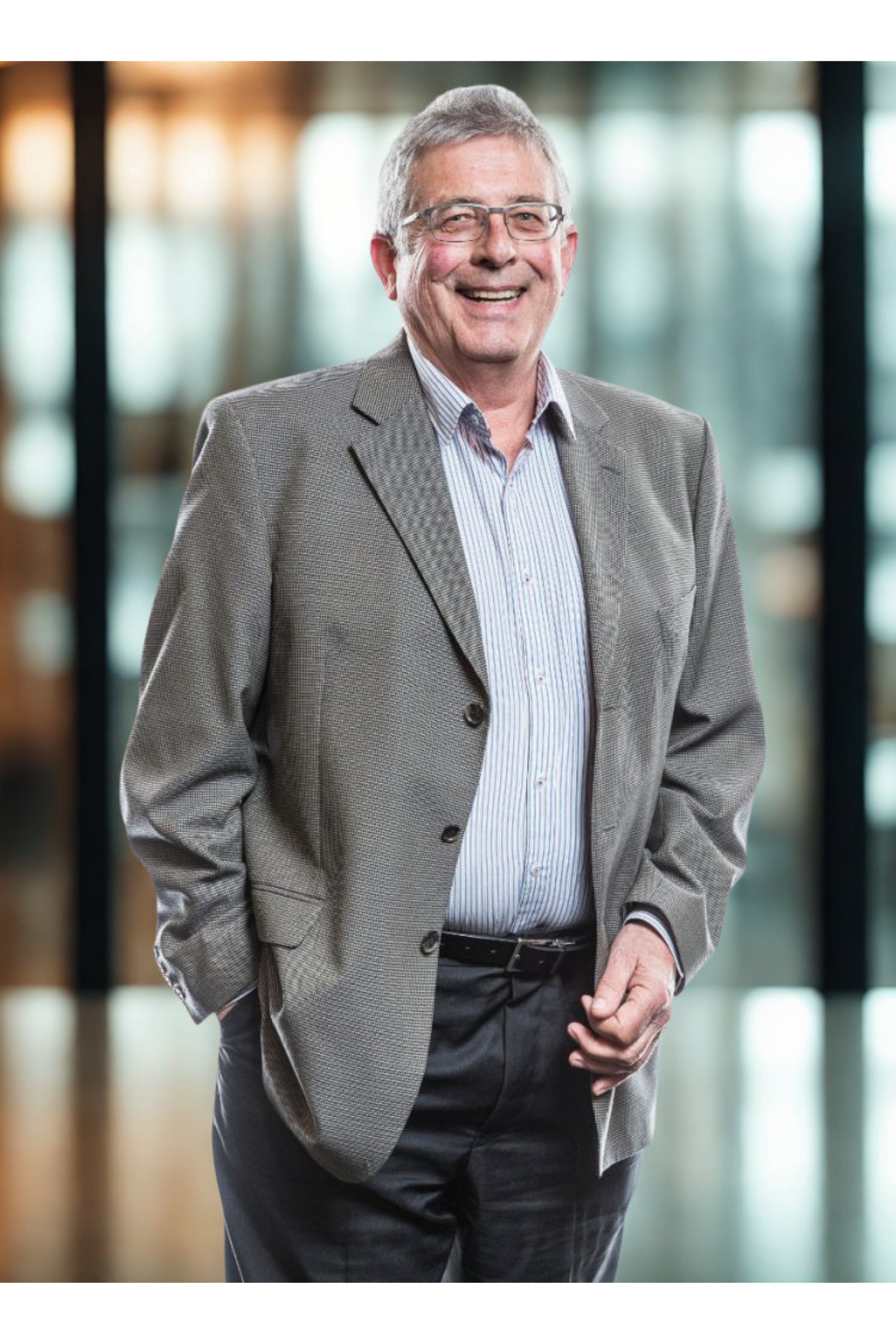 A smiling middle-aged man with gray hair and glasses, wearing a gray blazer and striped shirt, standing in a modern, brightly lit office or lobby area.