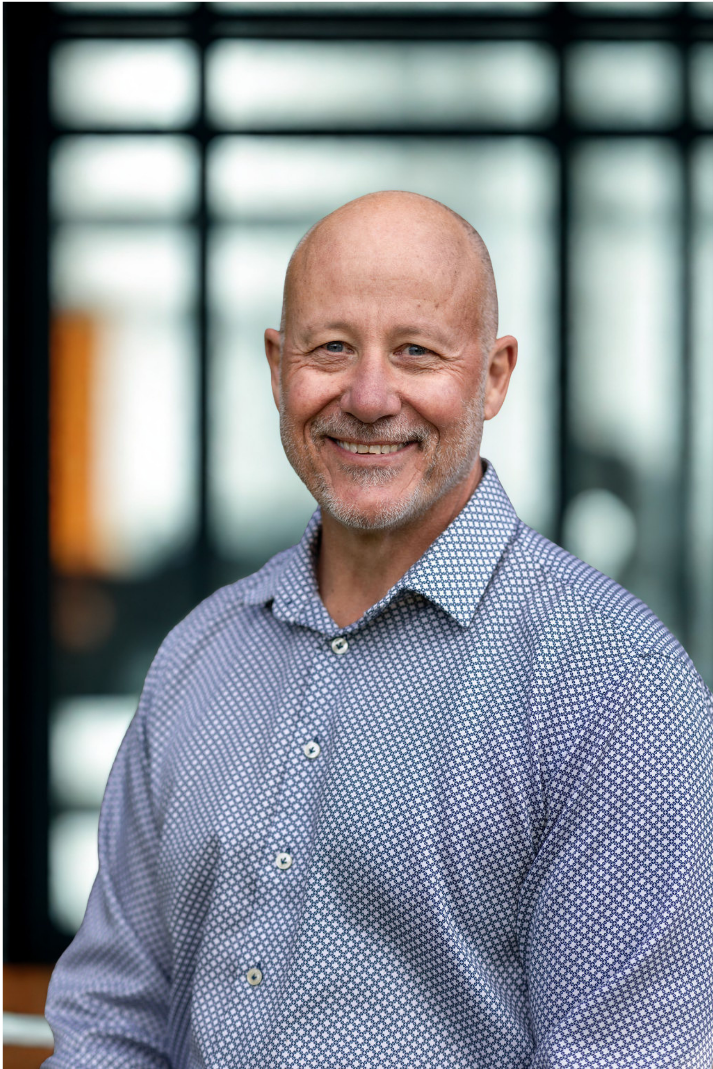 Close-up of a smiling middle-aged man with a bald head and a gray beard, wearing a blue and white patterned button-up shirt, standing indoors with blurred windows in the background.