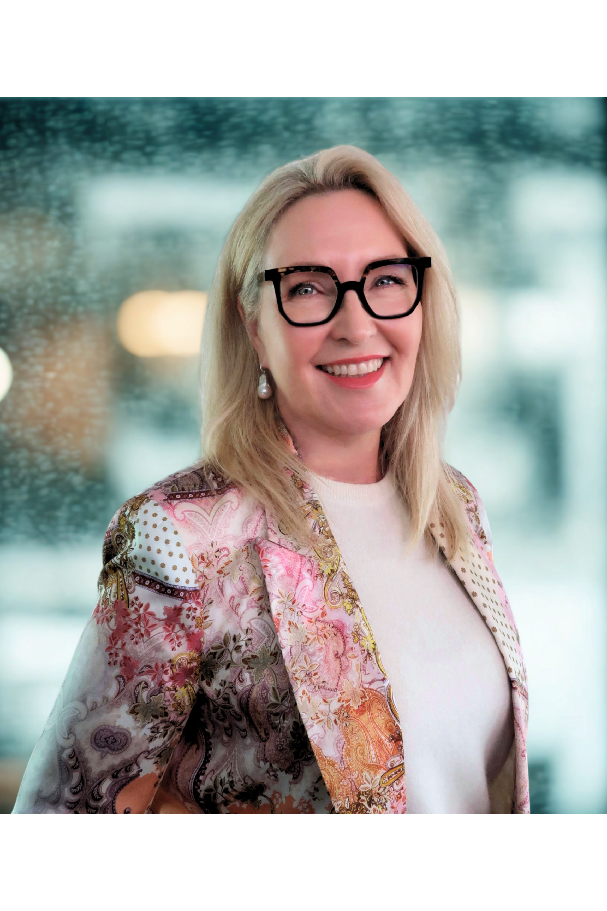 Woman with blonde hair, black glasses, white shirt, and a colorful patterned blazer, smiling in front of a blurred background of water and lights.